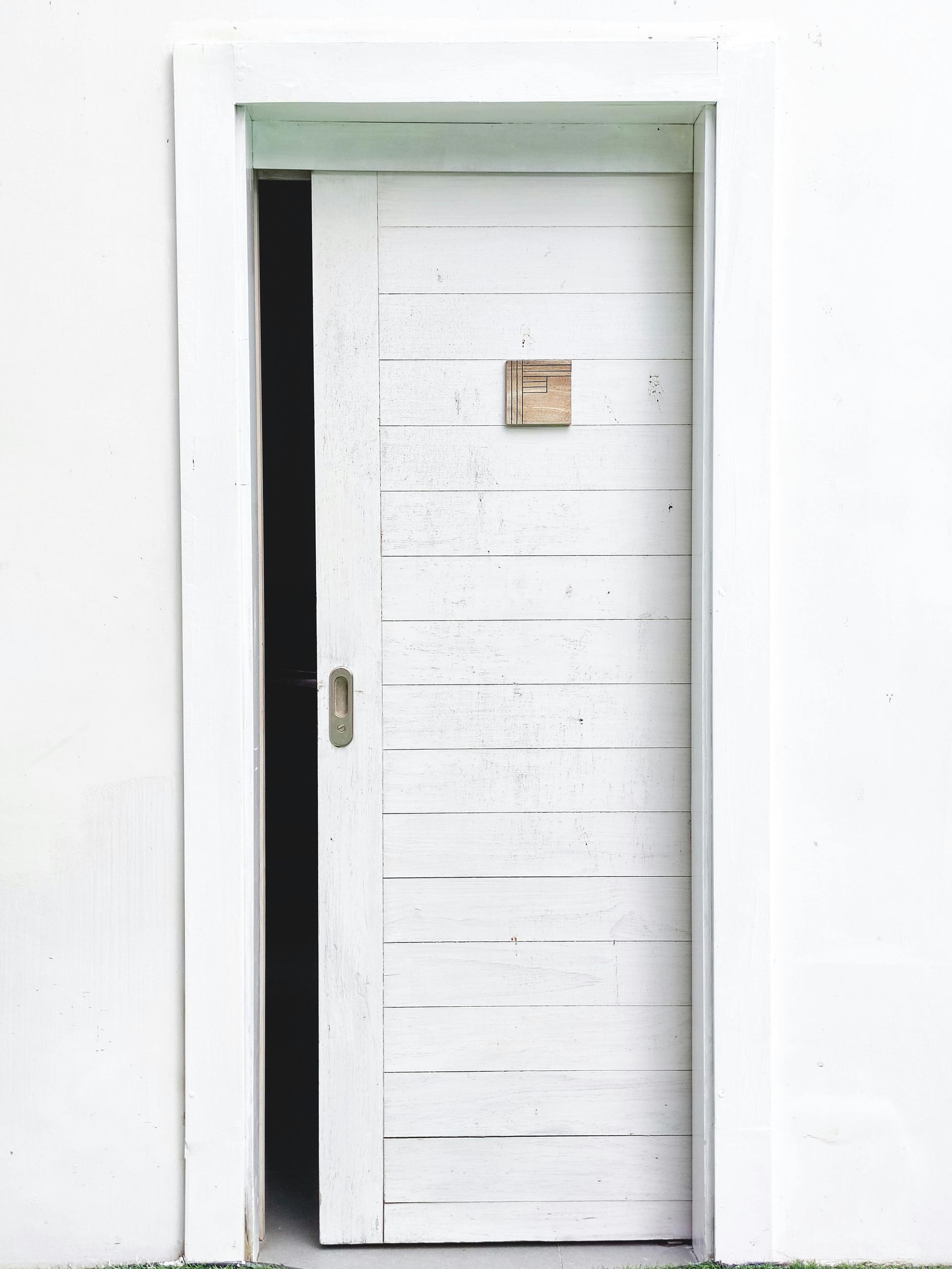 Porta bianca usurata leggermente socchiusa, inserita in uno stipite bianco contro una parete bianca.
