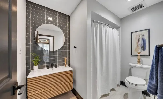 Modern bathroom with gray walls, black tile accent wall, white fixtures, and a wooden vanity.