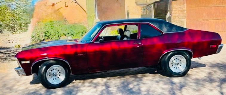 Red classic car parked on a sunlit street beside a beige wall with a shadowed background