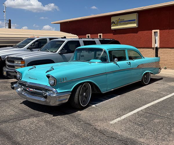 Turquoise vintage car parked in a lot beside a tan building under a blue sky.