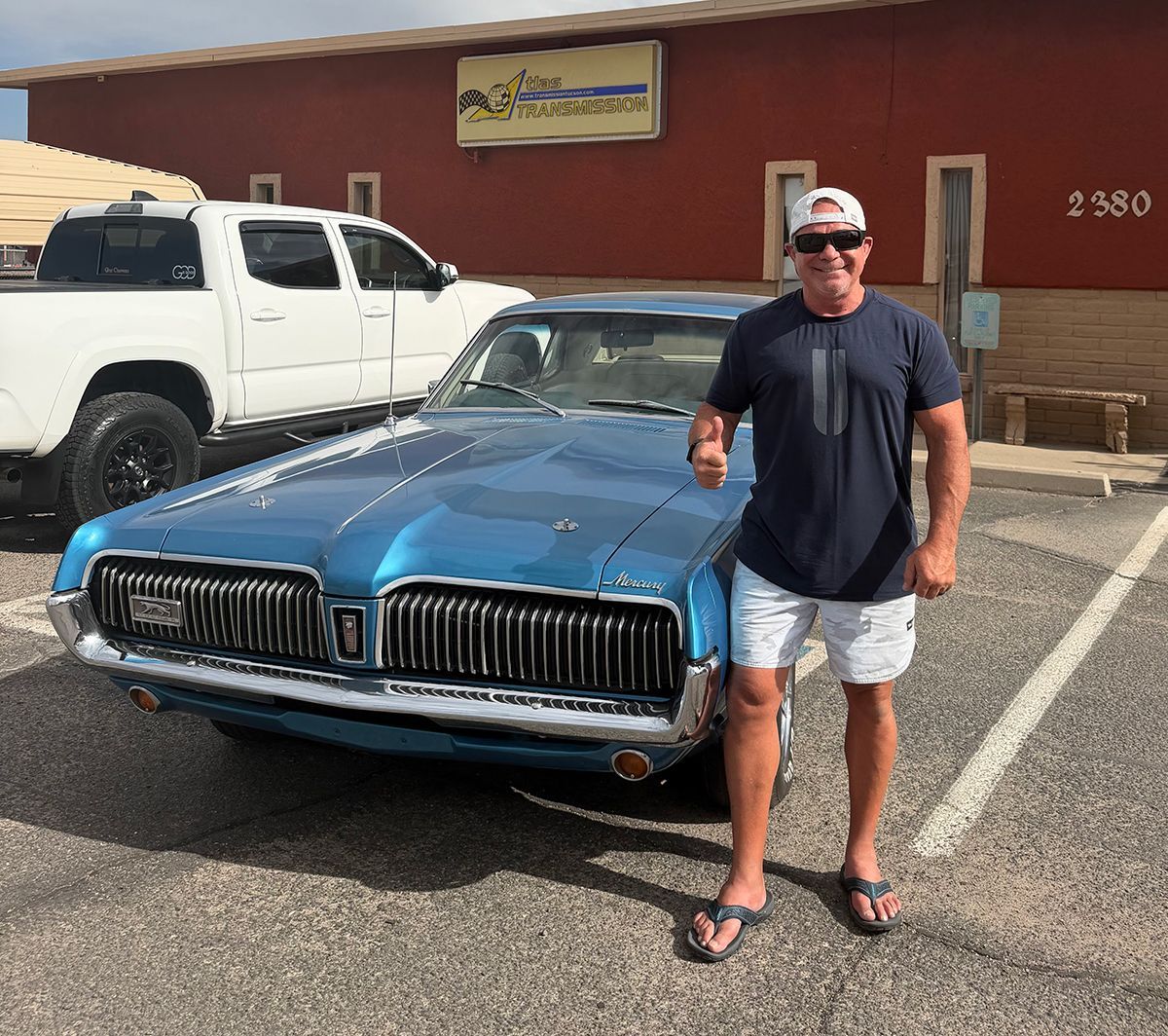 Man standing beside a blue classic car in a parking lot outside a building