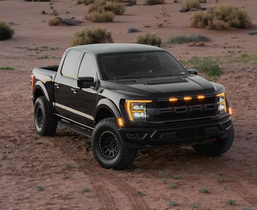 A black Ford F-150 Raptor pickup truck parked on a sandy desert terrain with illuminated front marker lights.