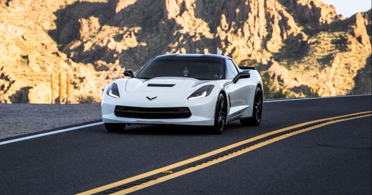 A white sports car is driving down a road with mountains in the background.