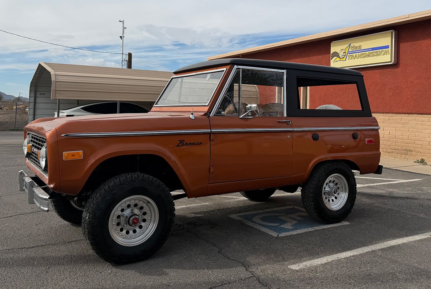 Brown vintage Ford Bronco parked outside a brick building with a black hardtop.