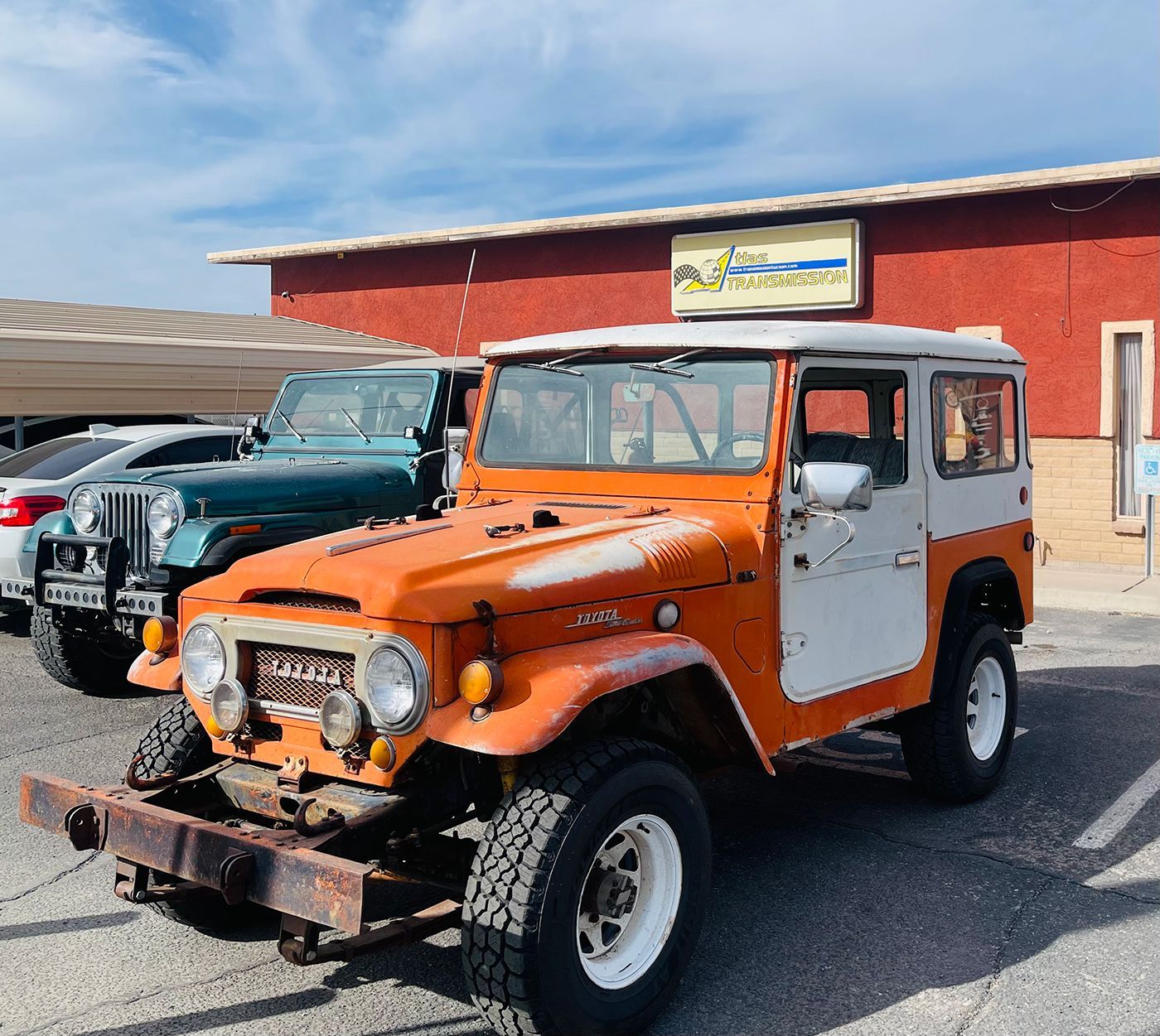 Orange and white vintage Toyota Land Cruiser parked in front of a building with a Jeep visible behind it.