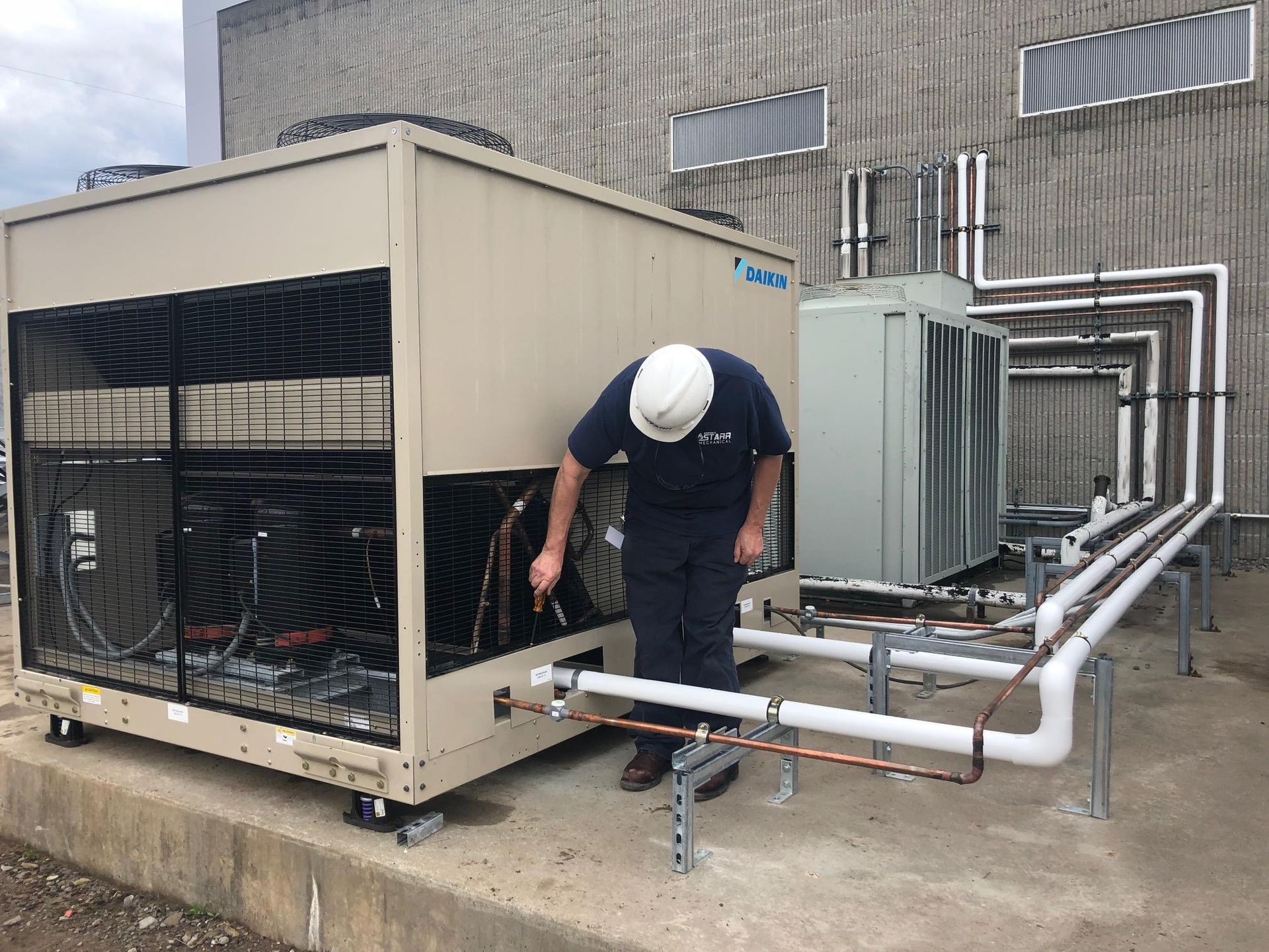 HVAC technician in a hard hat inspecting outdoor air conditioning unit with copper piping.
