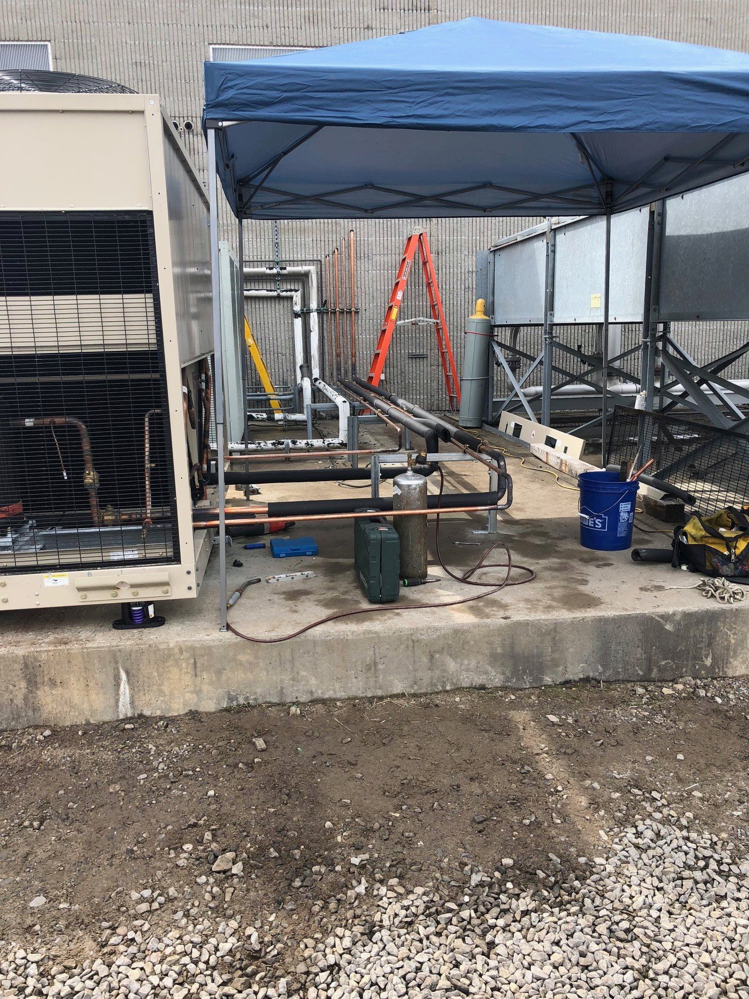 HVAC equipment under a blue canopy on a concrete pad. Tools, pipes, and a ladder are visible.