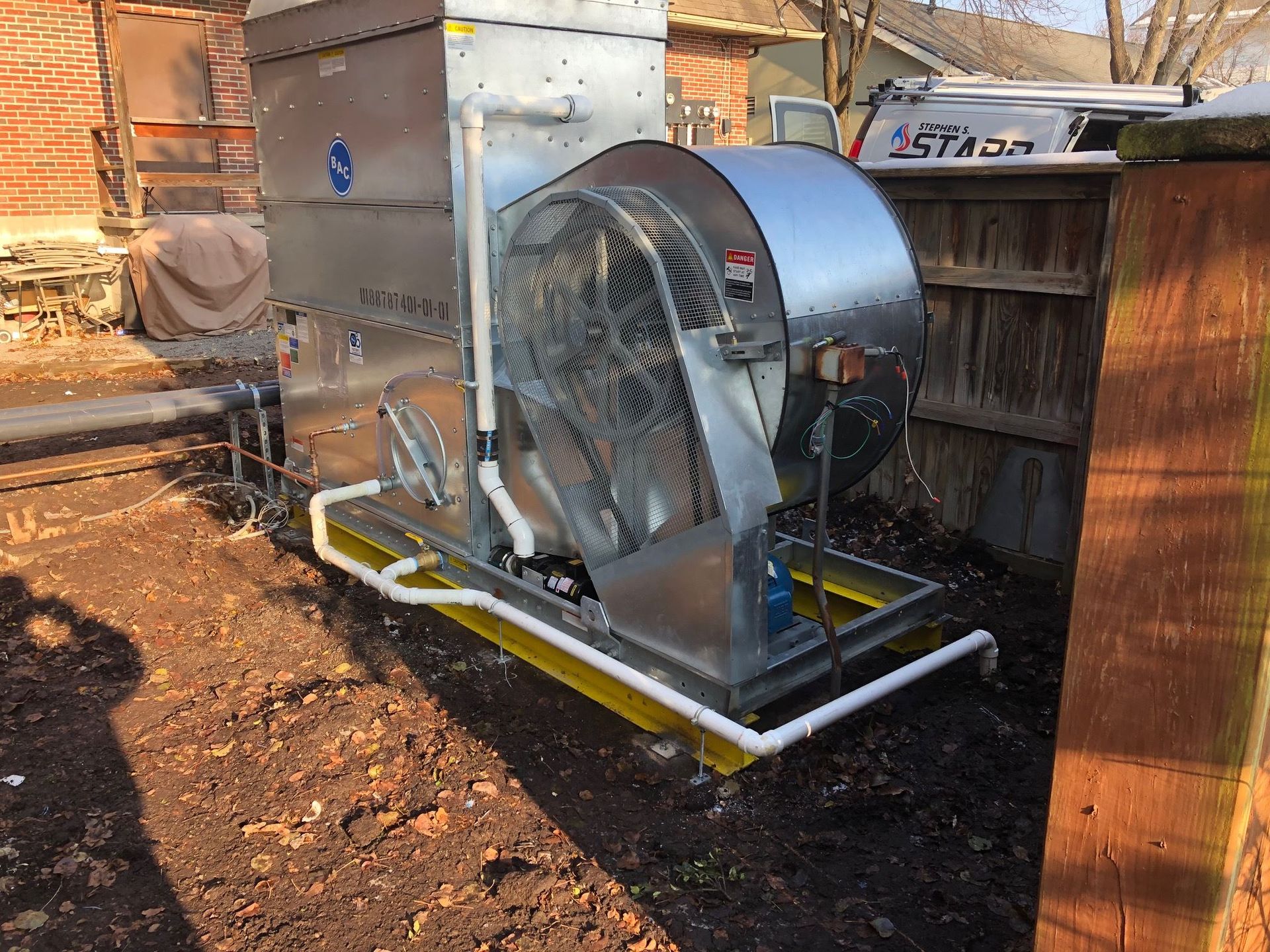 Large industrial fan and metal structure in an outdoor setting, on a yellow base, near a wooden fence.