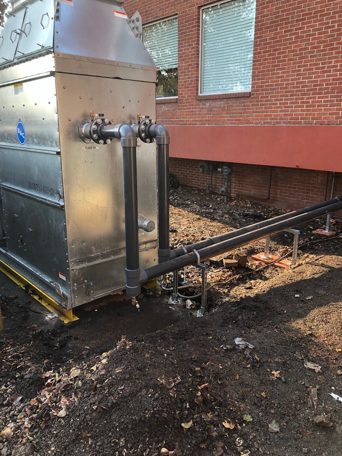 Metallic industrial unit with connected pipes near a brick building and dirt ground.
