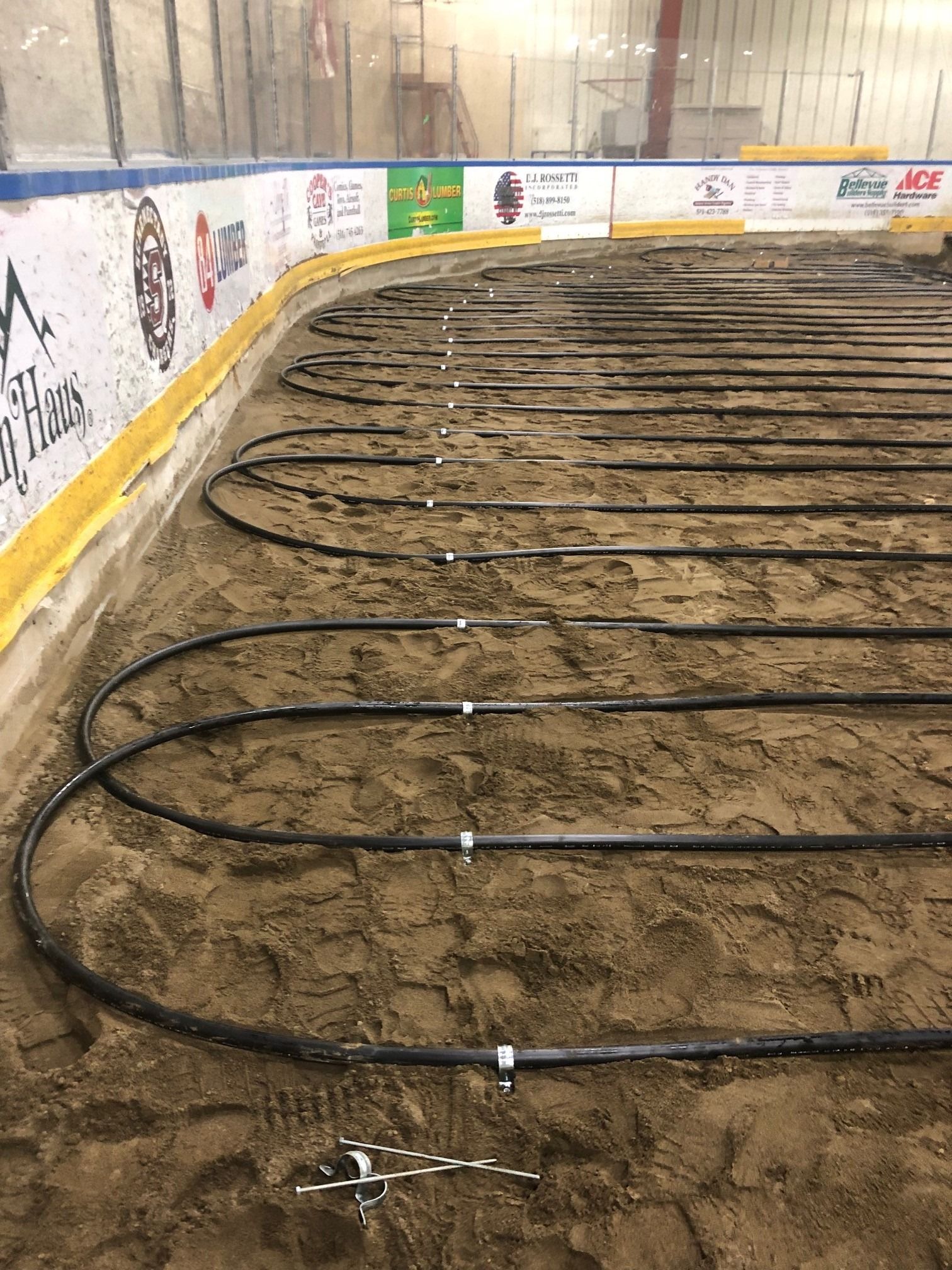 Pipes laid on sand for an ice rink floor, near the boards with advertising.