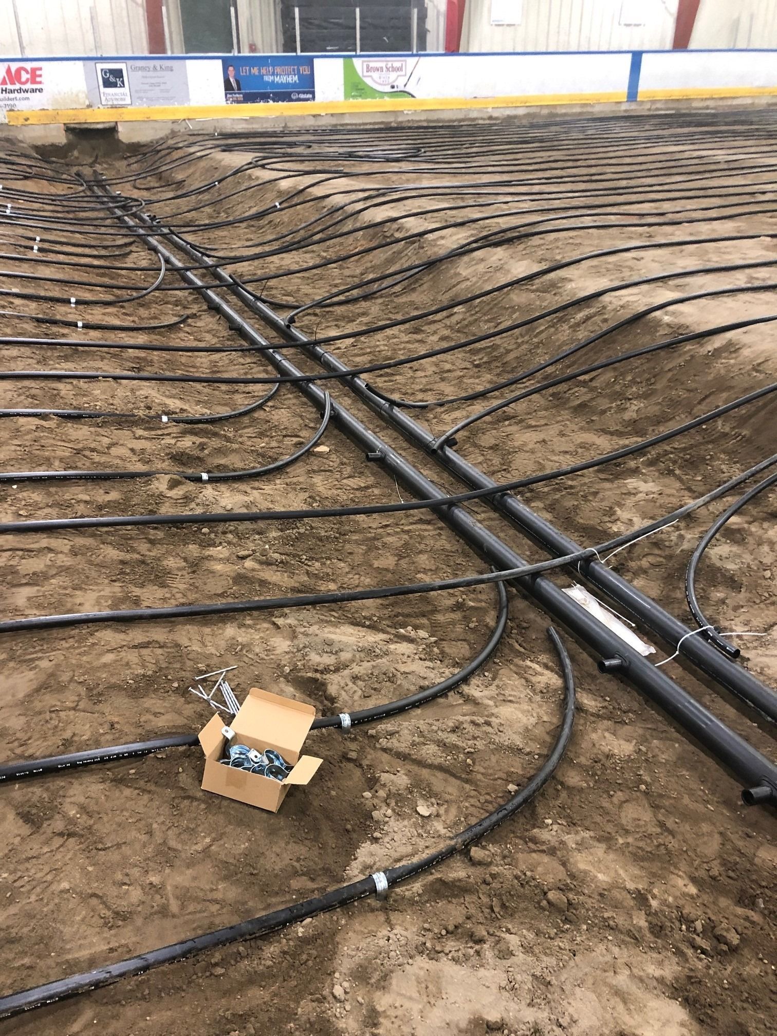 Pipes laid out on a dirt surface inside an arena, likely for an ice rink.