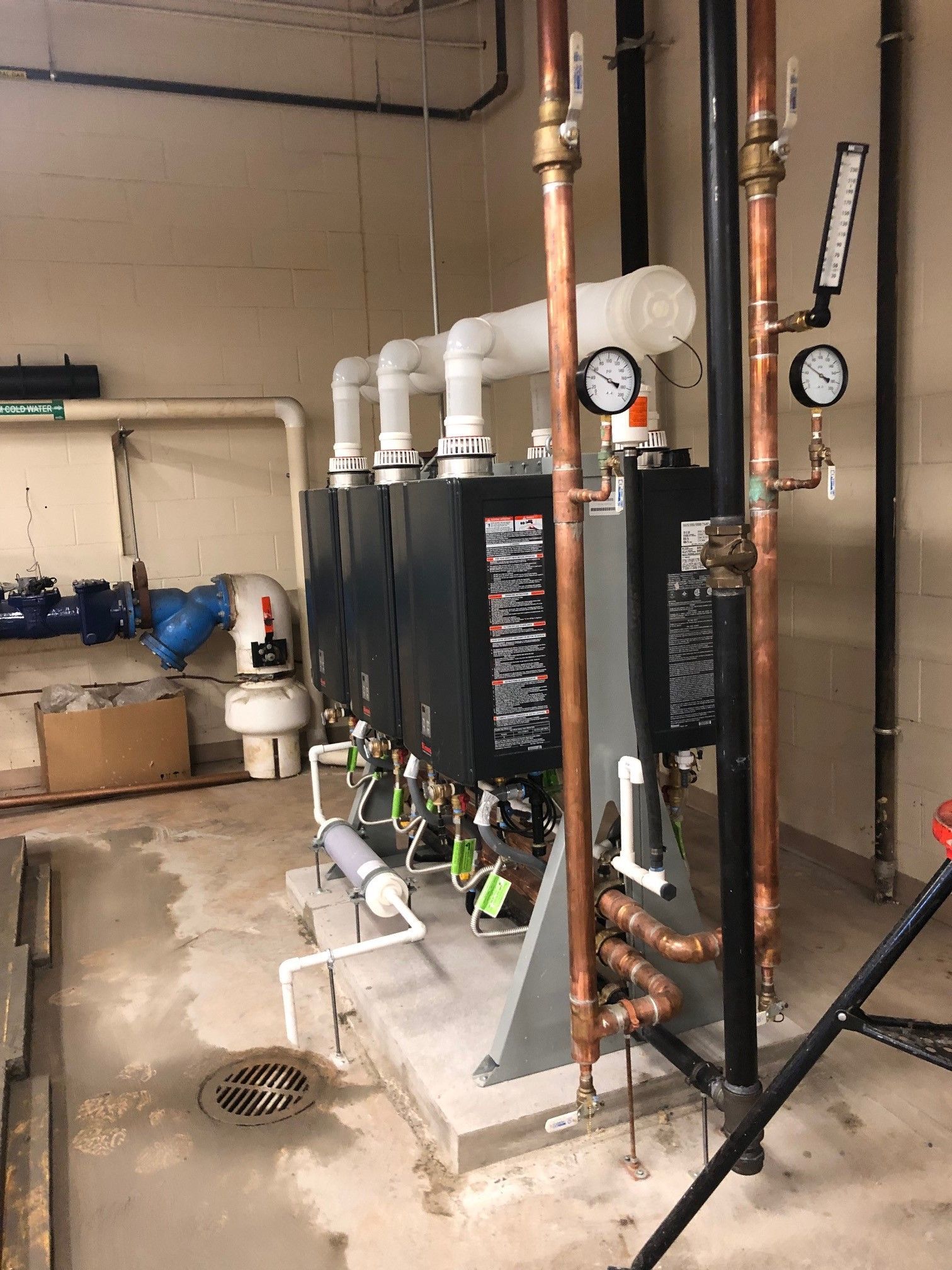 Row of black heating units with copper and white pipes in a utility room. Gauges and a drain are visible.