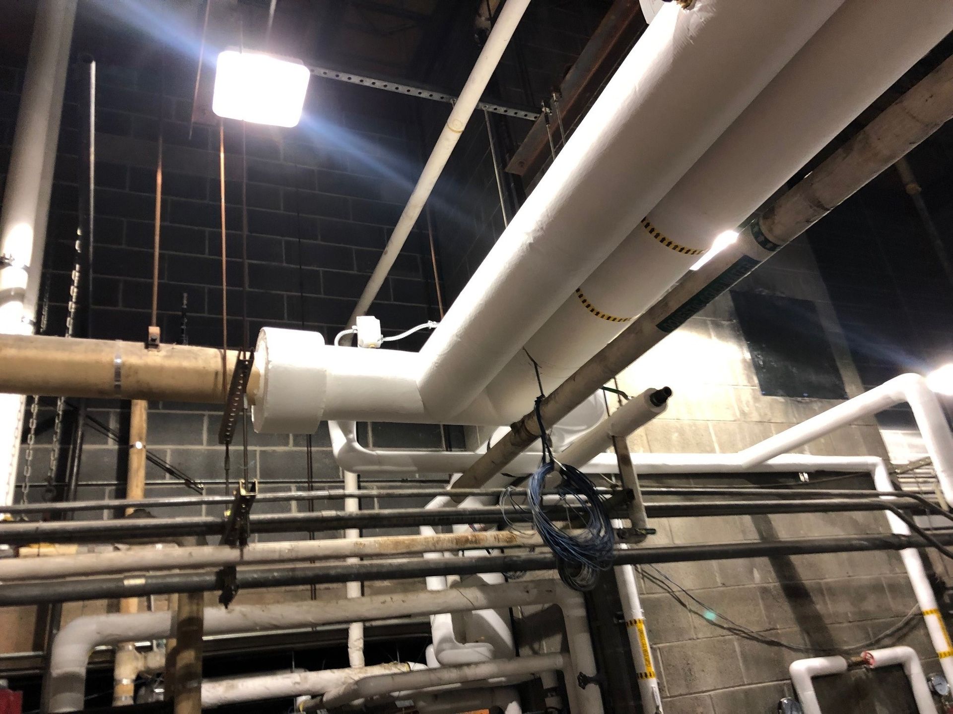 Pipes wrapped in white insulation run across a dark ceiling and brick wall. Some pipes are metal.
