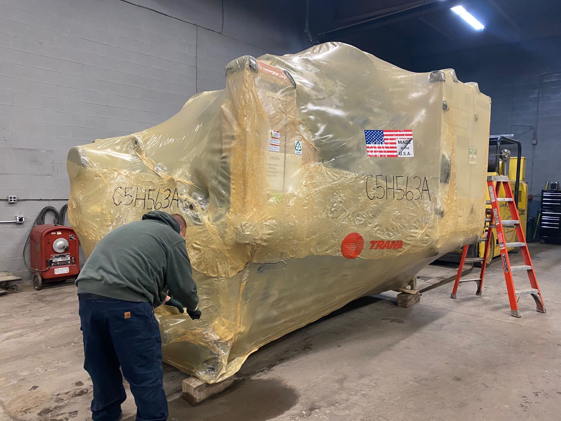 Man wrapping large machinery with clear plastic in a warehouse. A Trane label is visible.