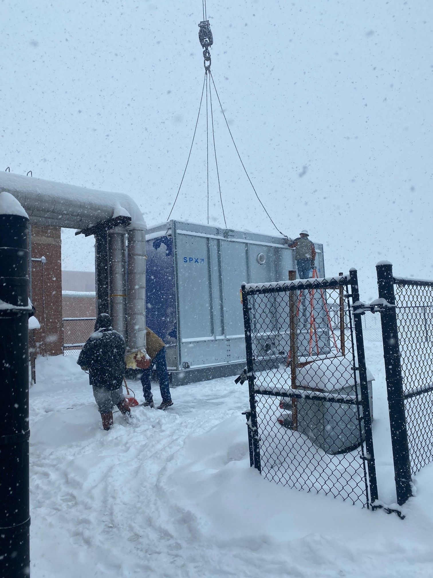 Workers installing a large, blue and silver unit outdoors in a snowy environment, crane overhead.