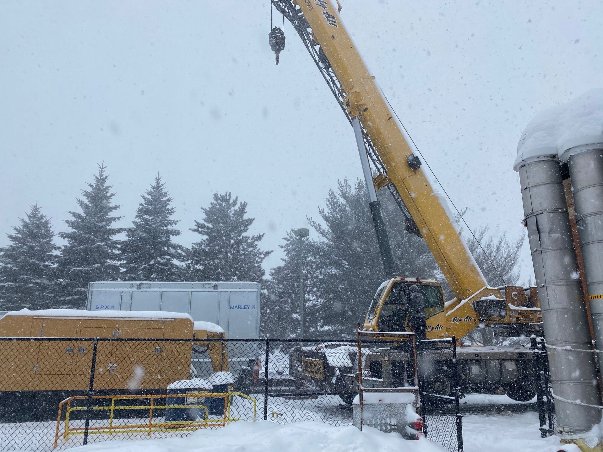 Yellow crane operating in a snowy setting with trees, a fence, and equipment.