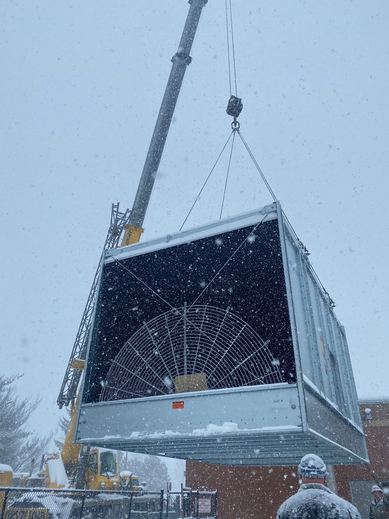 A crane lifting a large, square cooling tower in a snowy outdoor setting.