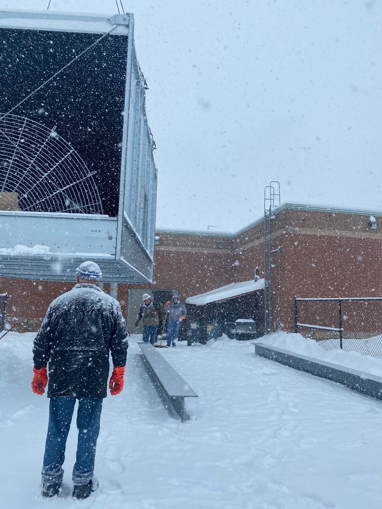 Man walks in snow towards a large metal structure, possibly a cooling tower, near a brick building.