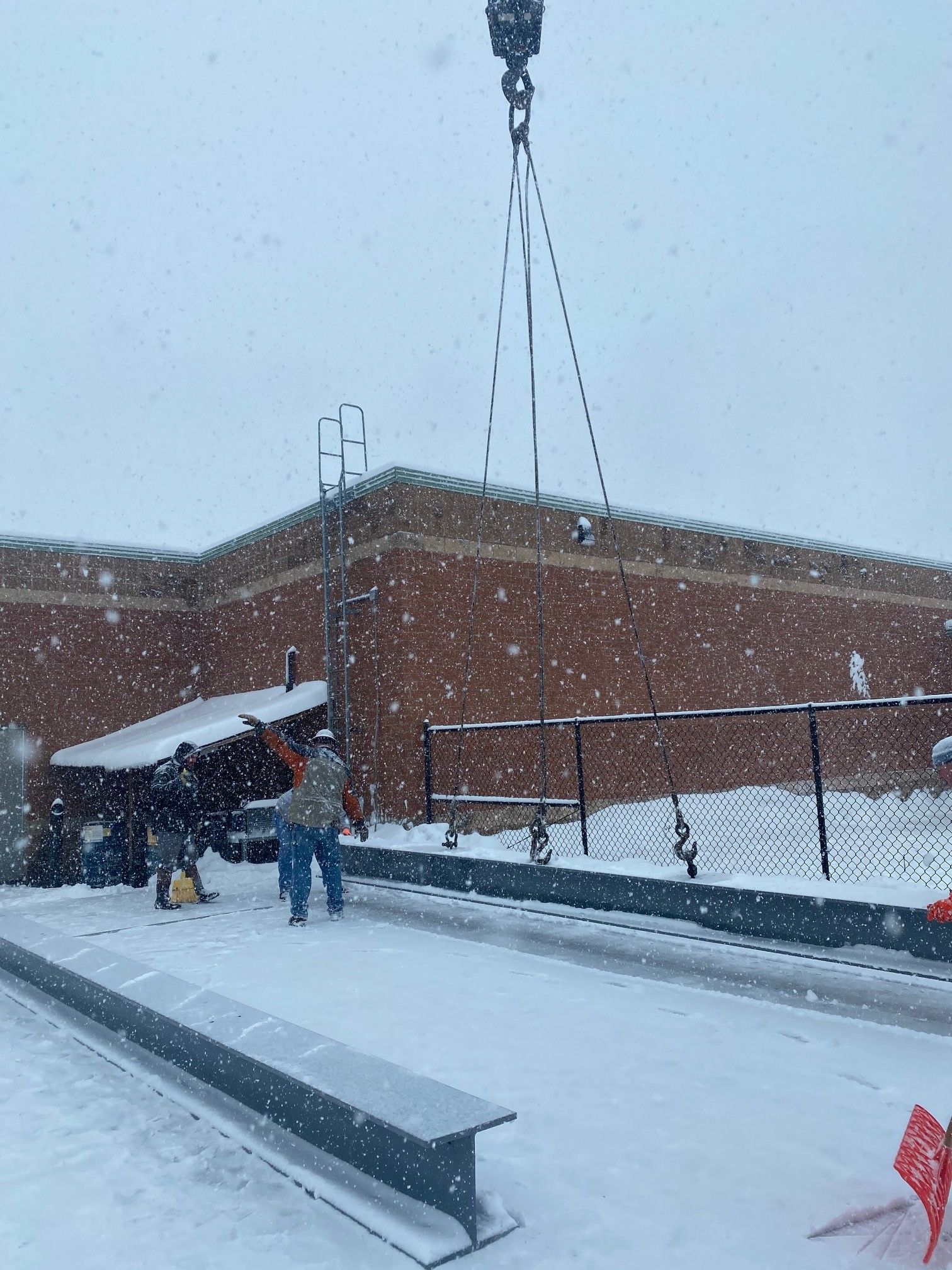 Crane lifting a steel beam on a snowy construction site. Workers in work clothes. Brick building in background.