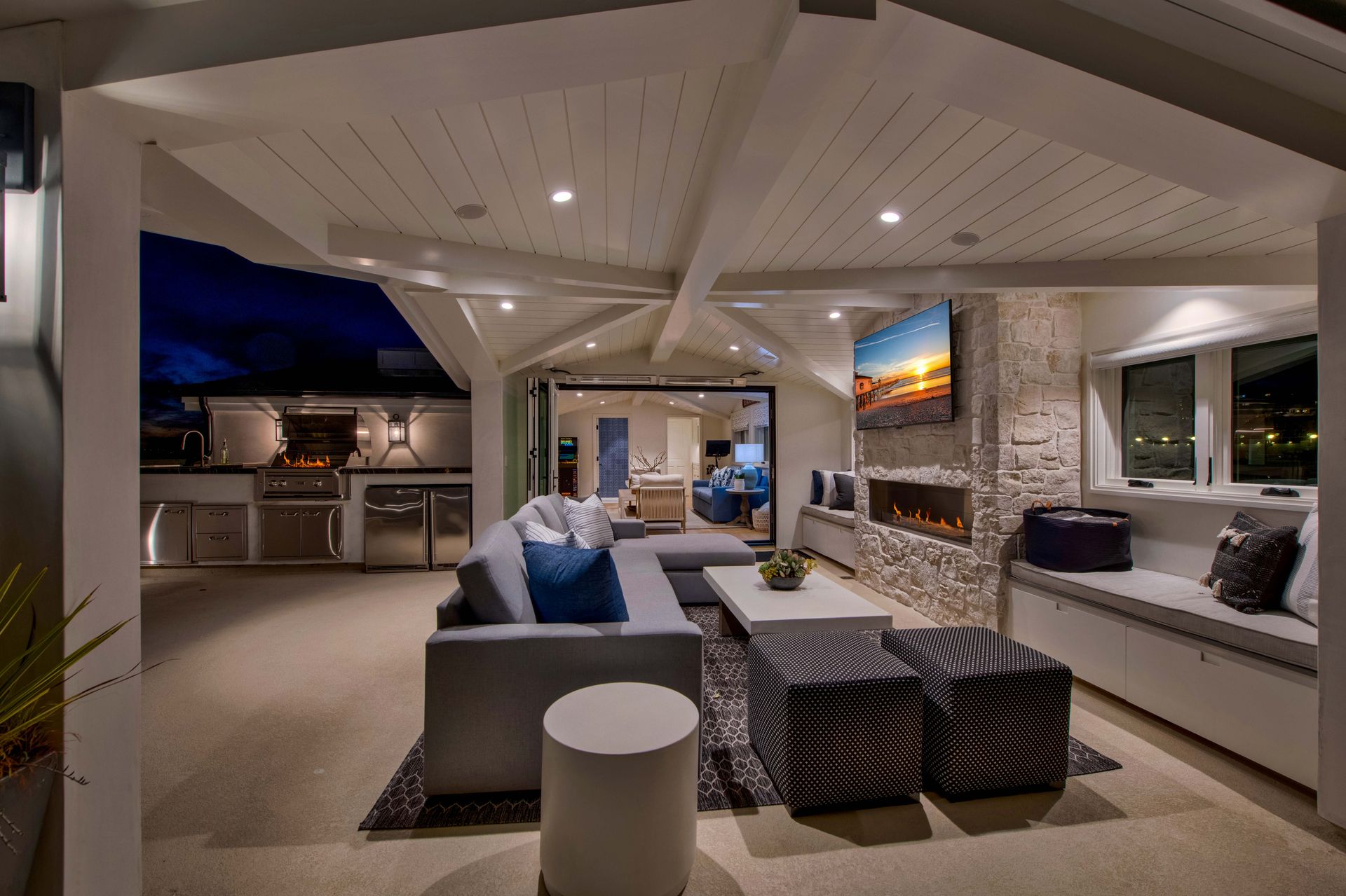 Luxury covered outdoor patio at night featuring a grey sofa, fireplace with a mounted TV, and stainless kitchen grill.