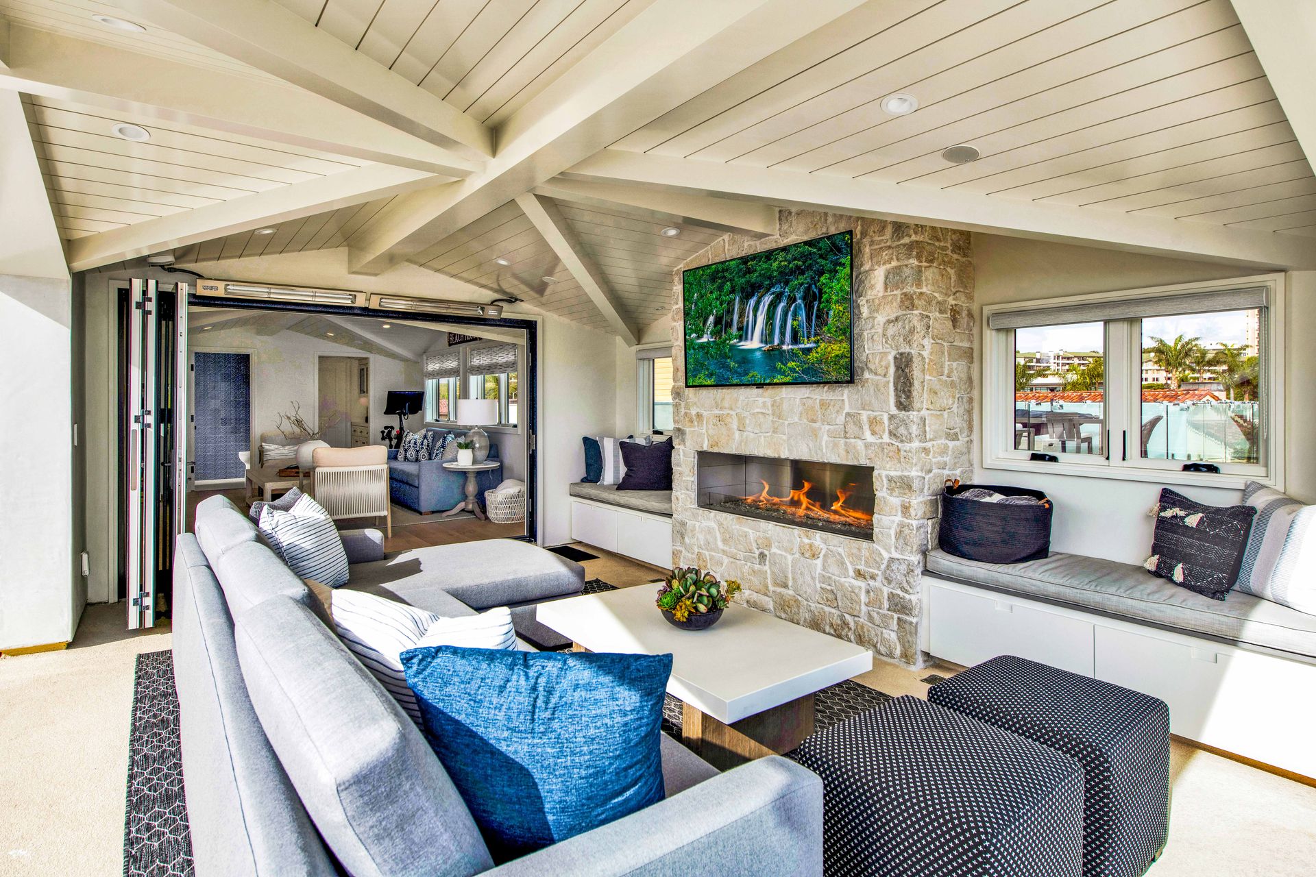 Modern living room with a stone fireplace, blue accent pillows, and folding glass doors leading to an adjacent room.
