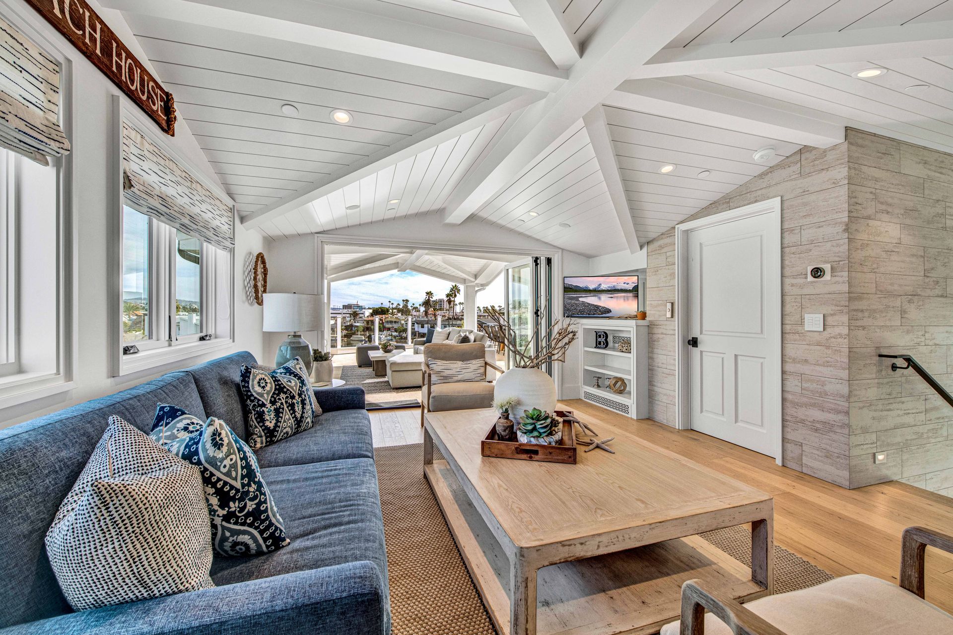 A bright coastal living room featuring a blue couch, wood coffee table, vaulted ceiling, and an open deck view.