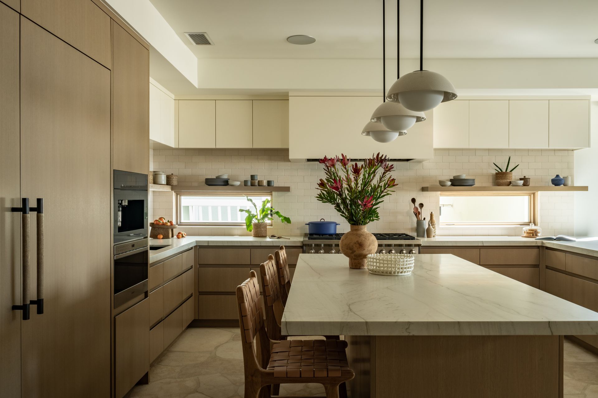 Modern kitchen with a marble-topped island, wooden cabinetry, cream-colored upper cabinets, and three hanging pendants.