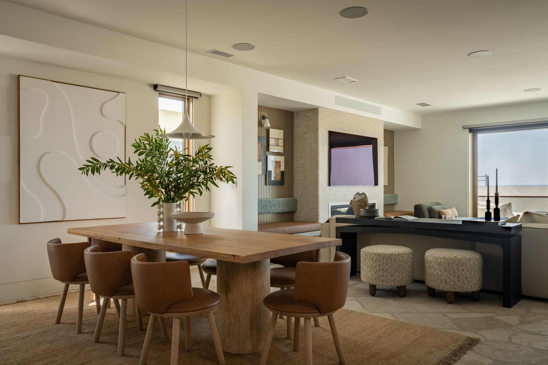 Dining area featuring a wooden table with tan chairs, textured wall art, and a lounge space with a TV and stool seating.