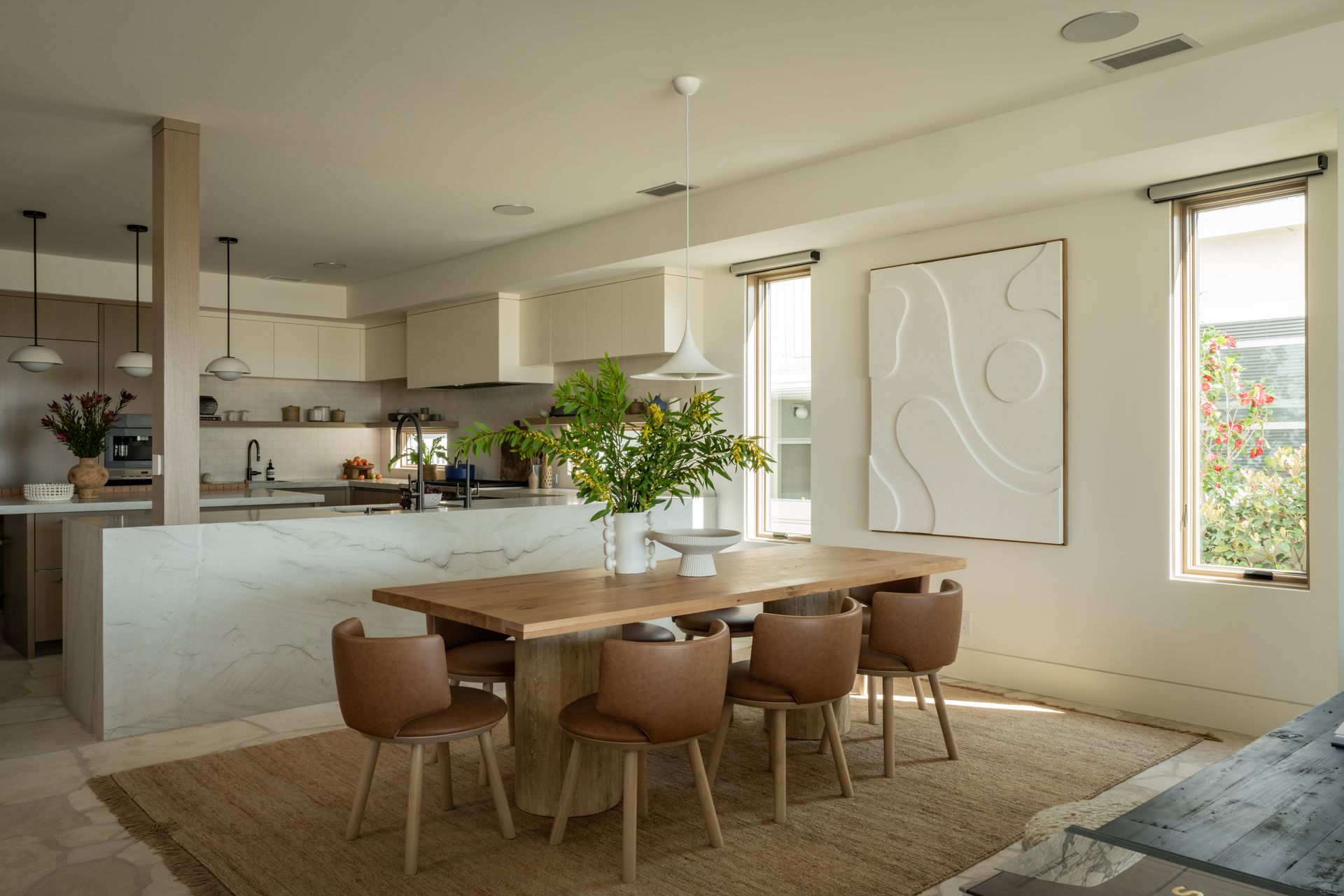 Modern kitchen with light wood cabinets, marble island, and wooden dining table with six brown chairs on a jute rug.