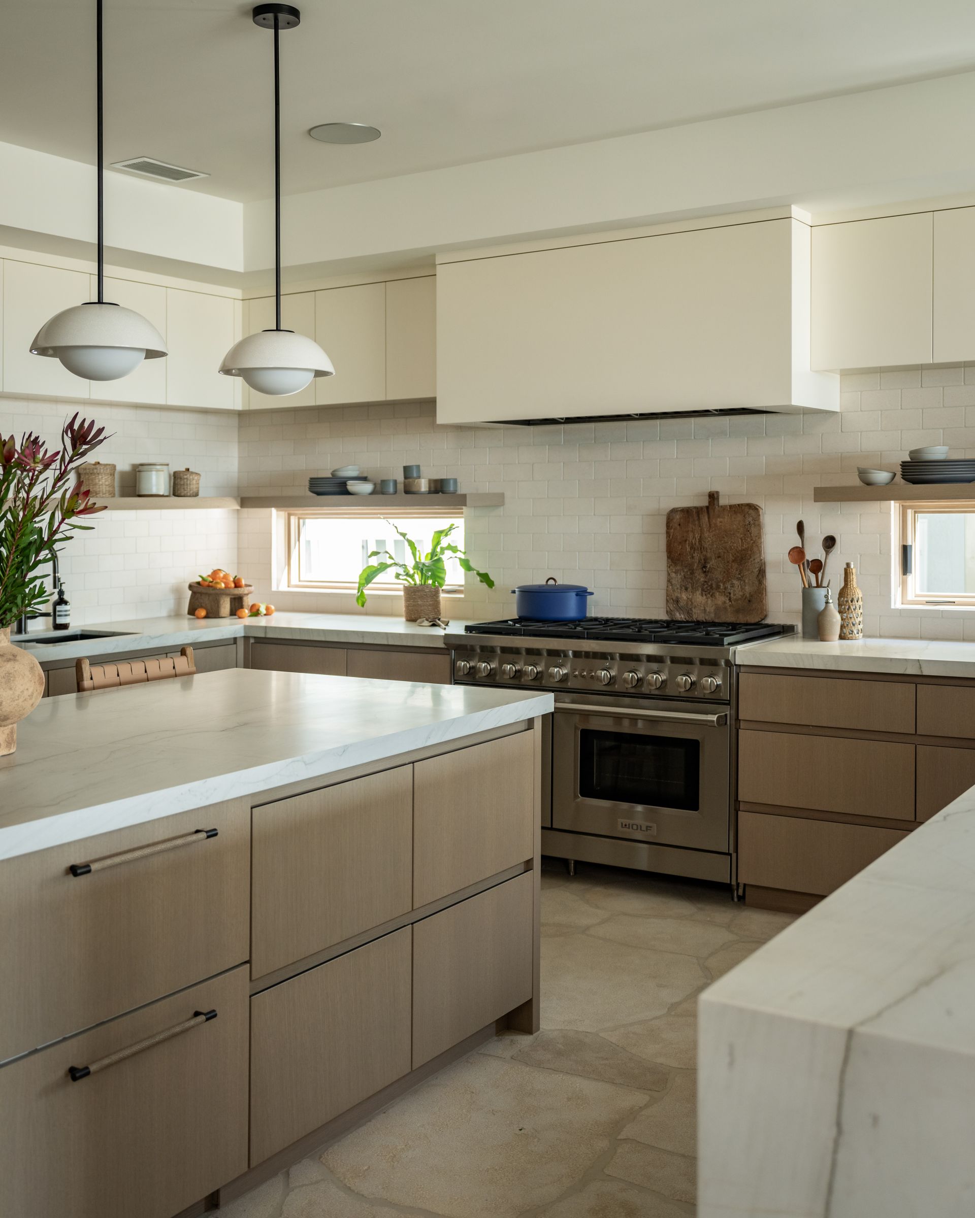 A modern kitchen with light wood cabinetry, stone countertops, a stainless steel range, and two white pendant lights.