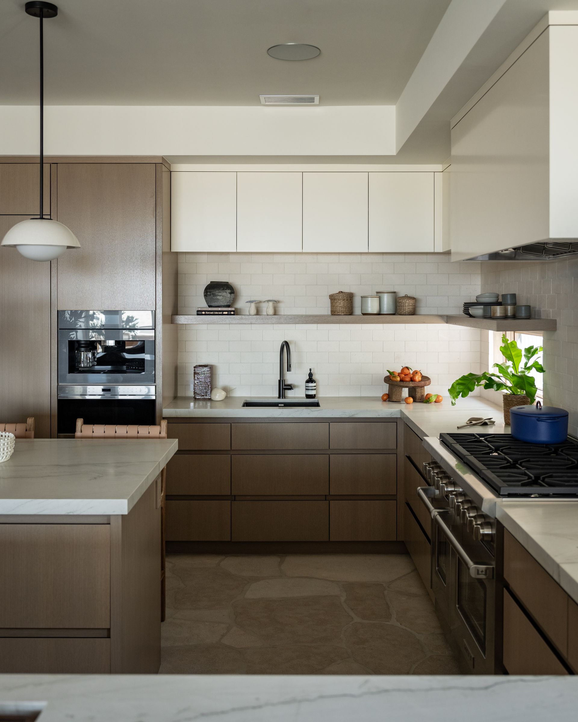 A modern, two-toned kitchen features taupe cabinets, white upper cabinets, light stone flooring, and a stone backsplash.