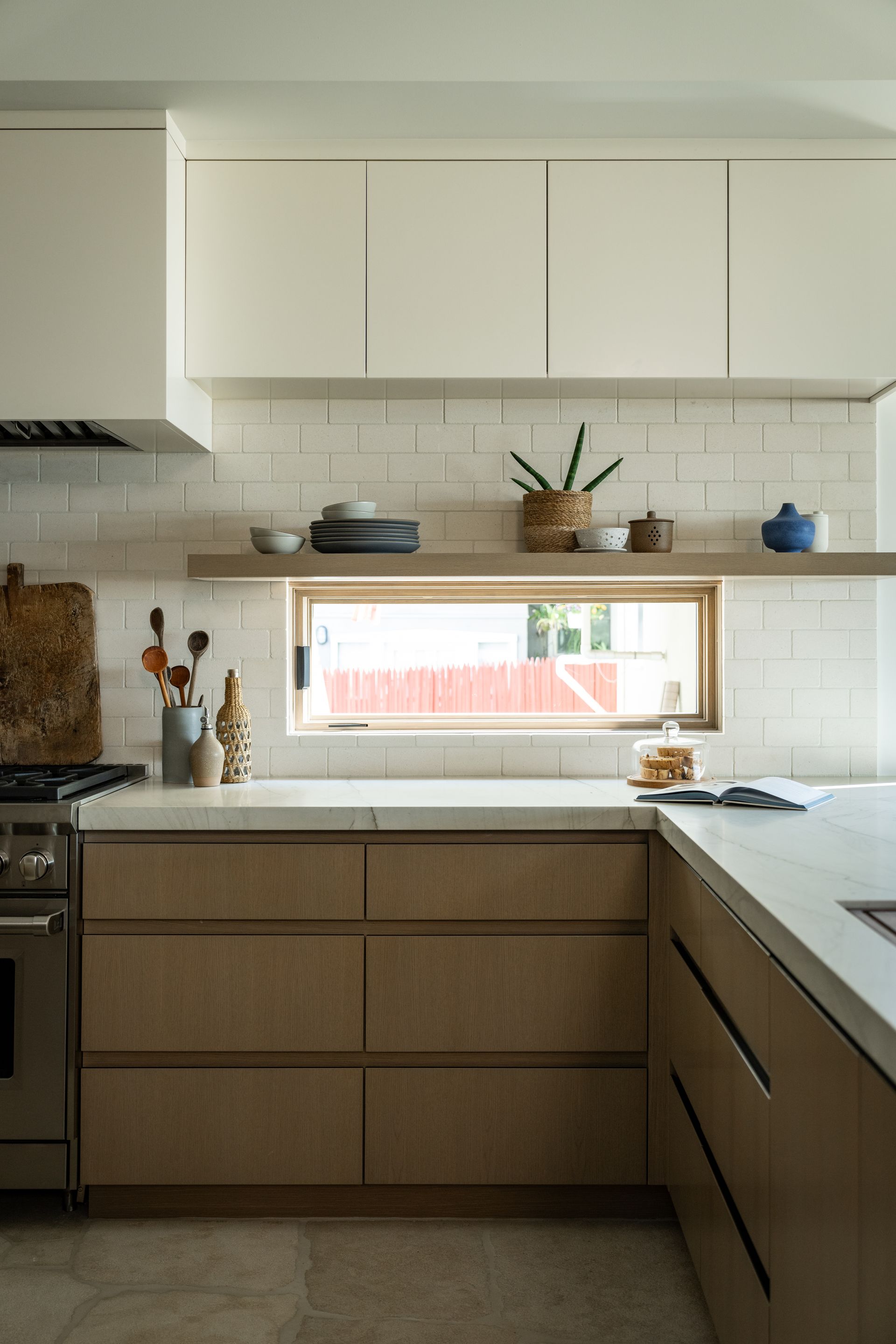 A modern kitchen with light wood lower cabinets, white upper cabinets, a tiled backsplash, and a window above the counter.