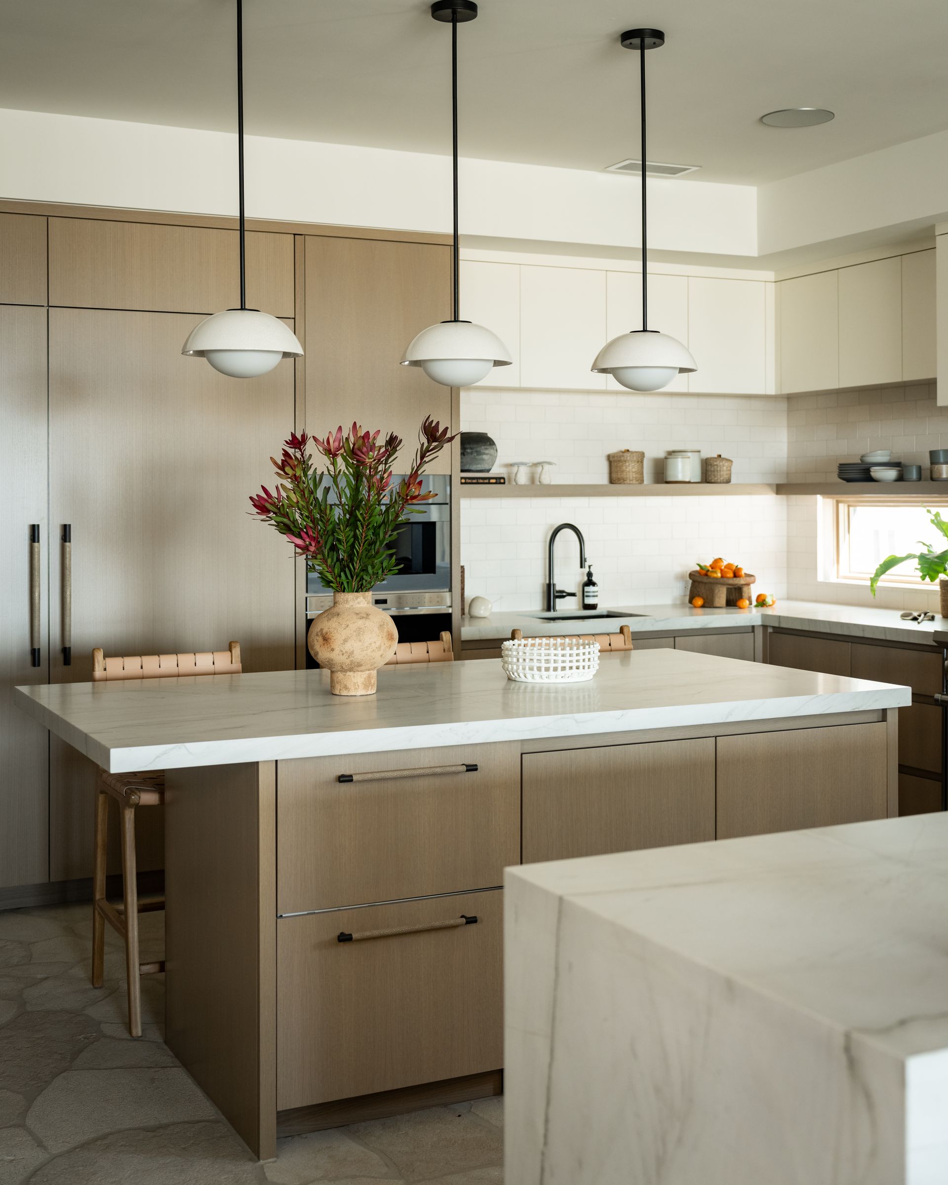 A modern kitchen with light wood cabinetry, a white stone island, three pendant lights, and a vase of flowers.