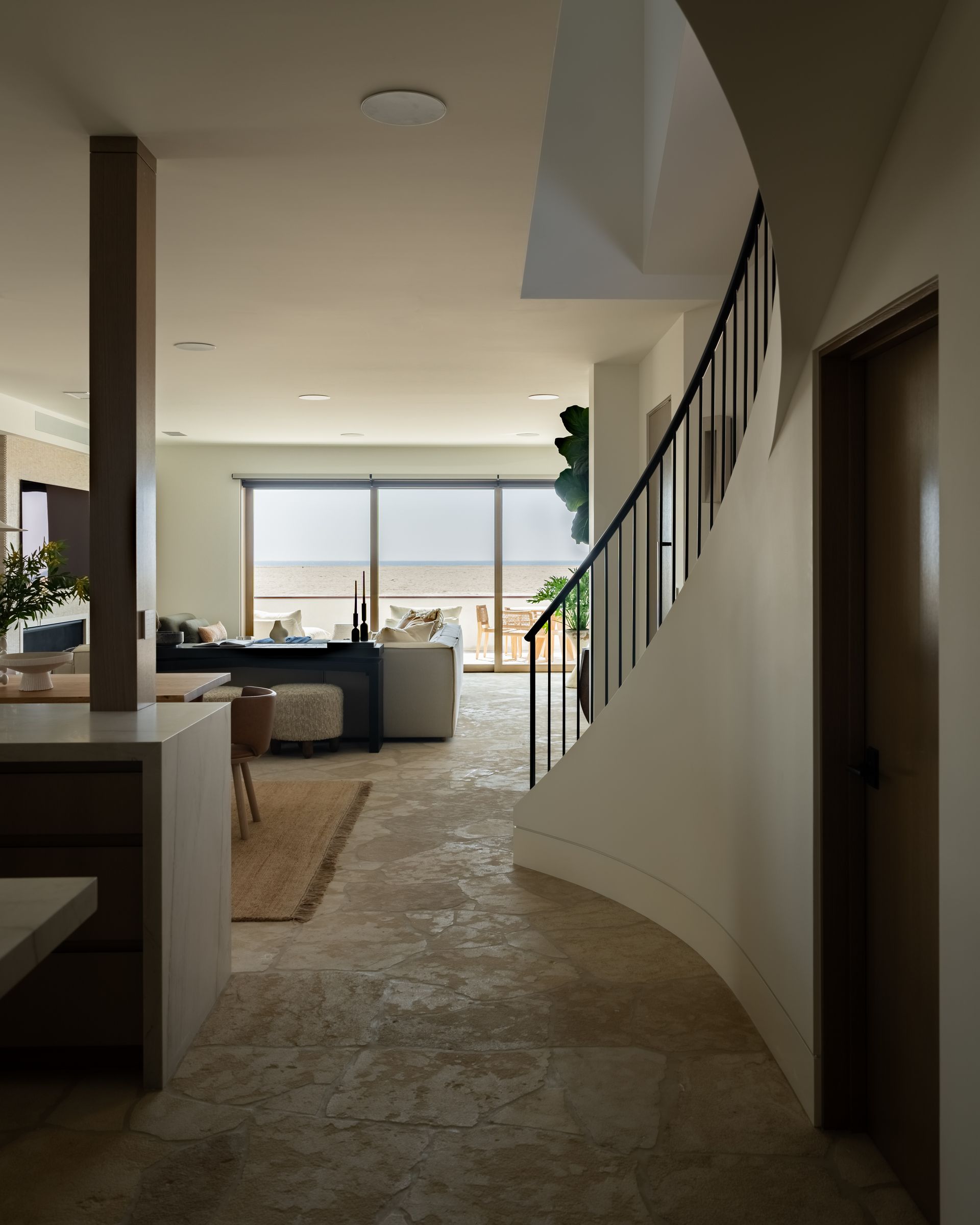 A hallway with textured stone floors leads to a bright living area, framed by a curved staircase with a metal railing.