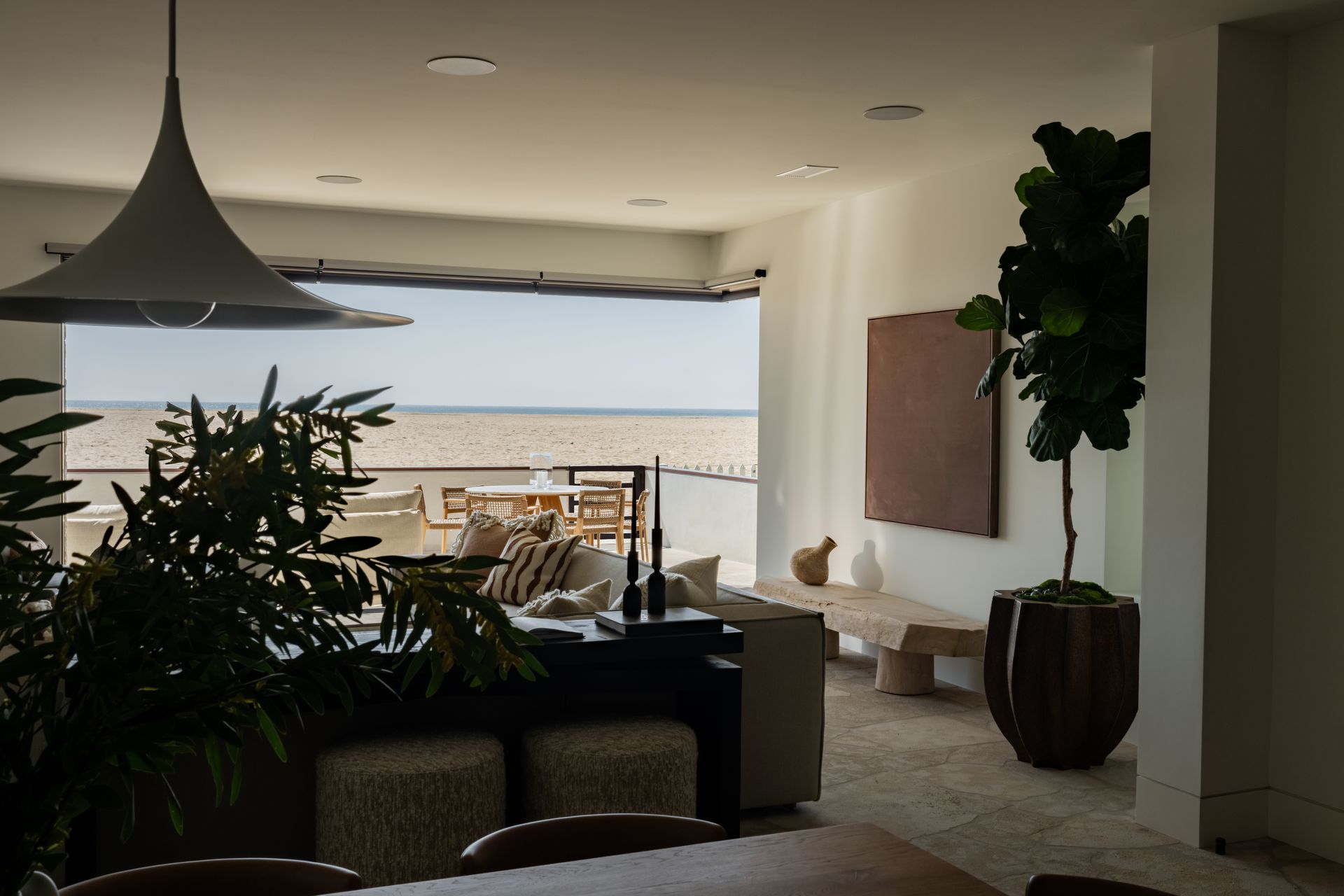 A modern living room opens onto a beach view, featuring a large potted plant, minimalist furniture, and a pendant lamp.