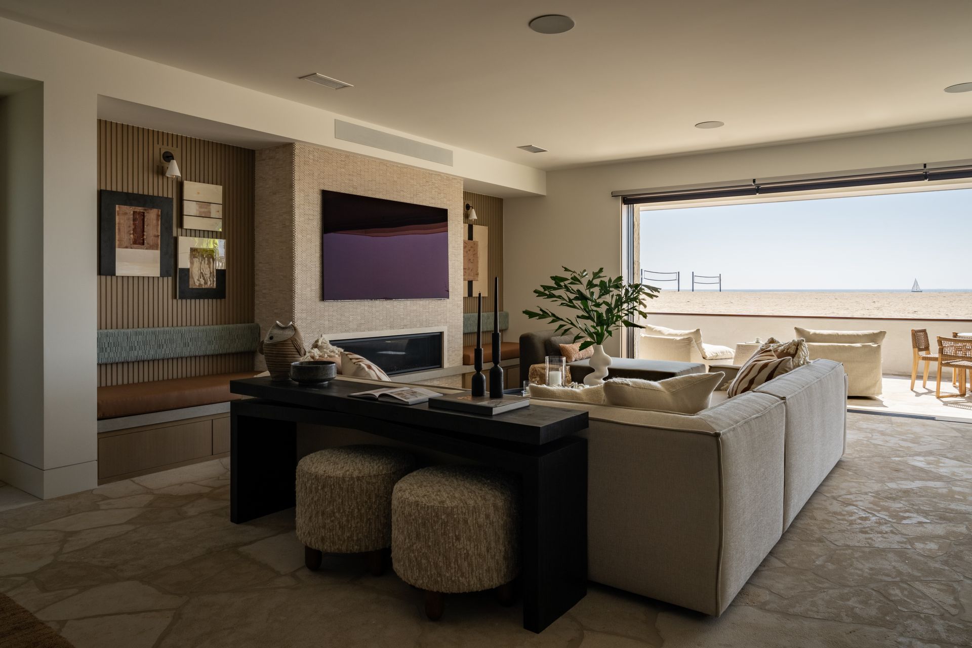 A modern living room with a large sofa, textured stools, a fireplace, and a wall of windows looking out onto a terrace.