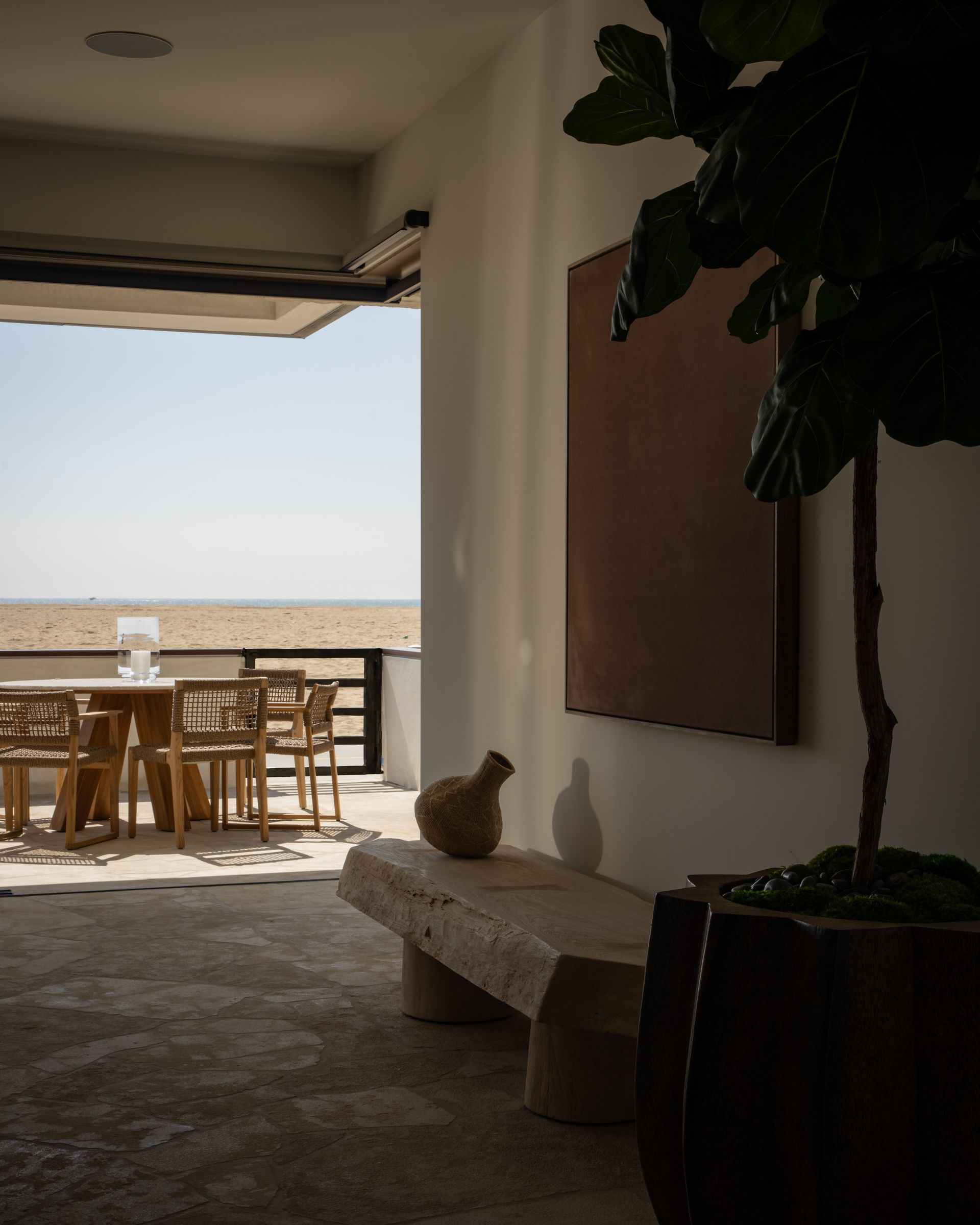A dimly lit entryway with a stone bench and large potted plant leading to a sunlit patio with outdoor dining.