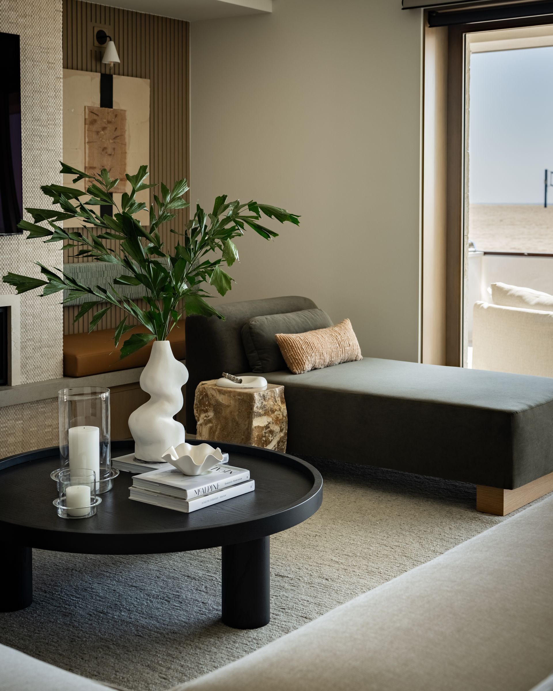 A modern living room with a dark round coffee table, a green plant in a white vase, and a grey chaise lounge near a window.