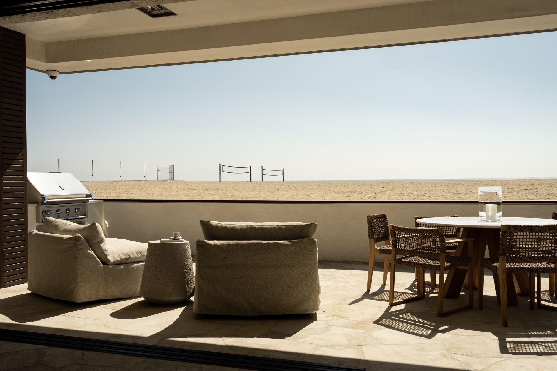 A sunlit patio with beige lounge chairs, a dining set, and a grill, overlooking a sandy beach with volleyball nets.