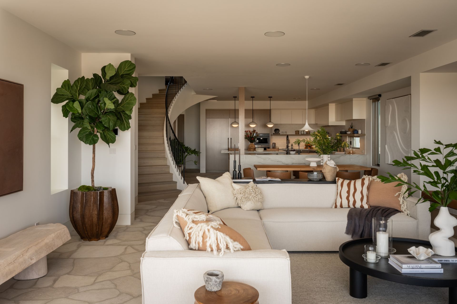 A modern, neutral-toned living room featuring a large beige sectional sofa, a curved staircase, and indoor plants.