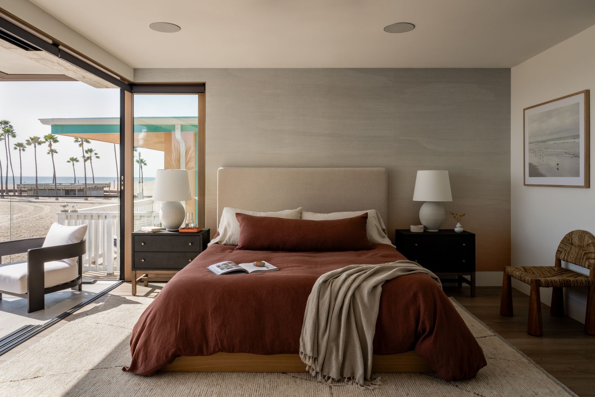 A modern beachside bedroom features a platform bed with deep red bedding, flanked by black nightstands and white lamps.