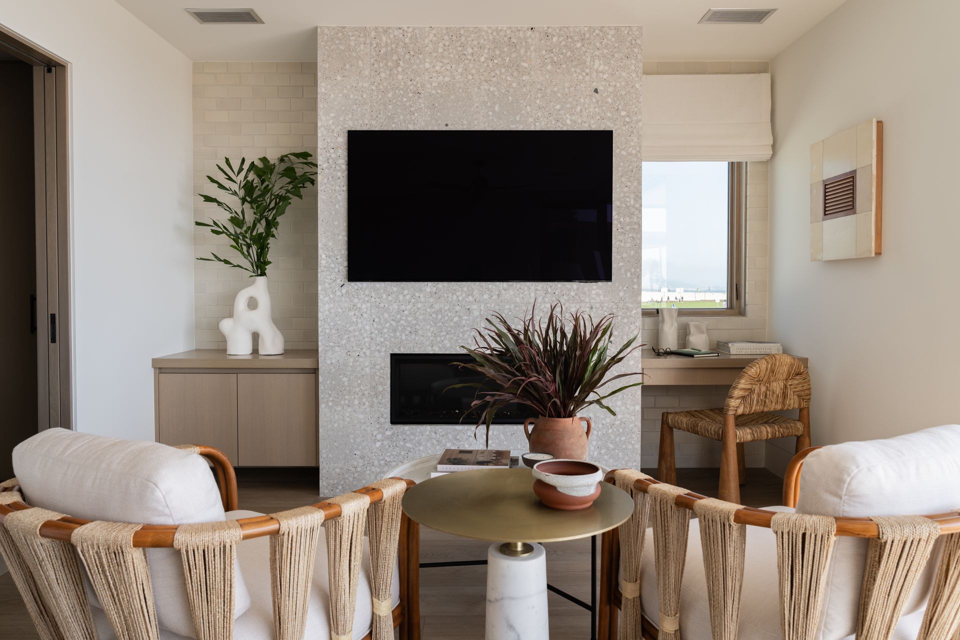 Modern living room with a terrazzo fireplace, two woven chairs, a round brass coffee table, and a small built-in desk.