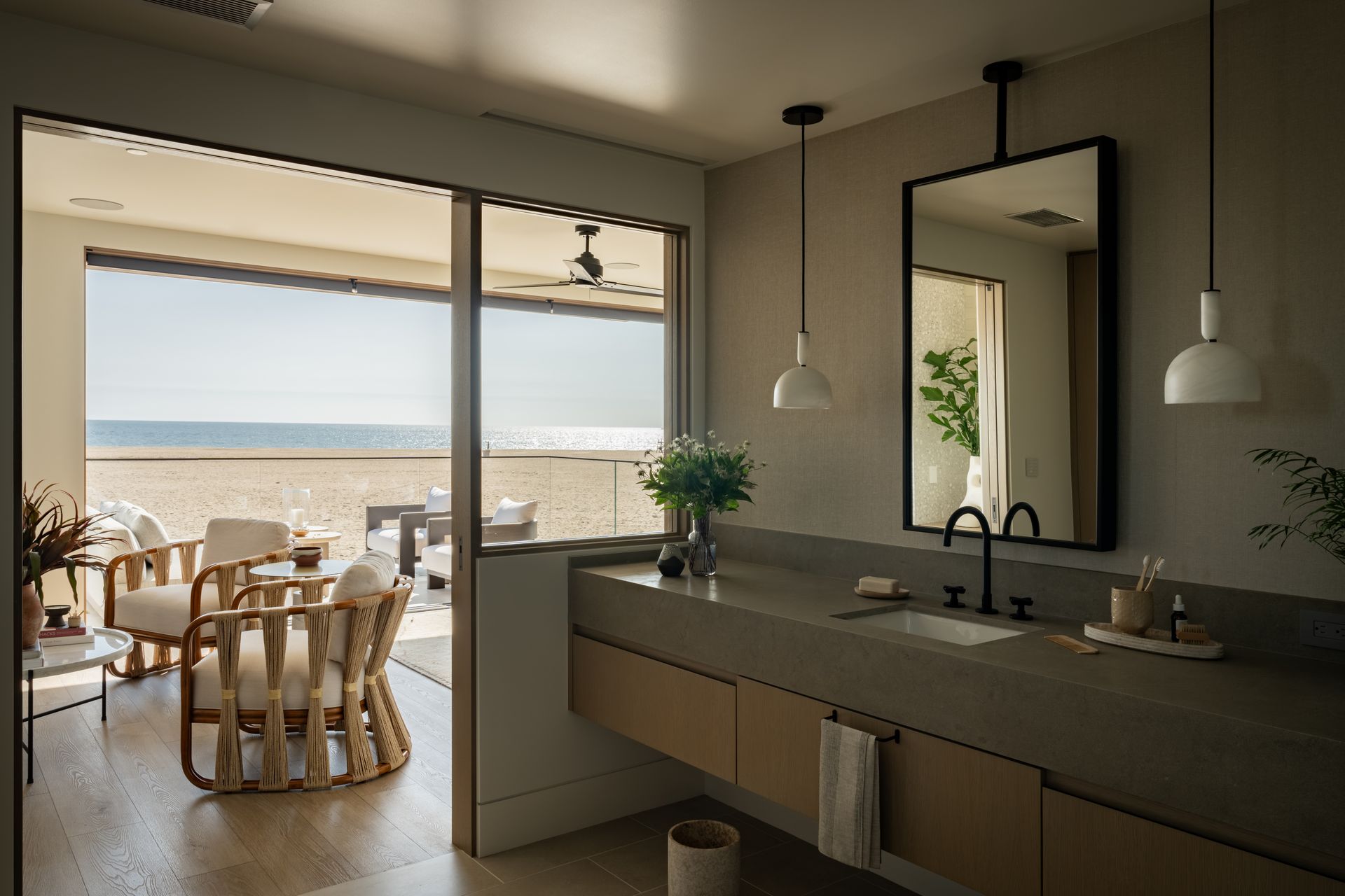 Modern bathroom with vanity and mirror overlooking a sunlit beach with outdoor seating through large glass doors.