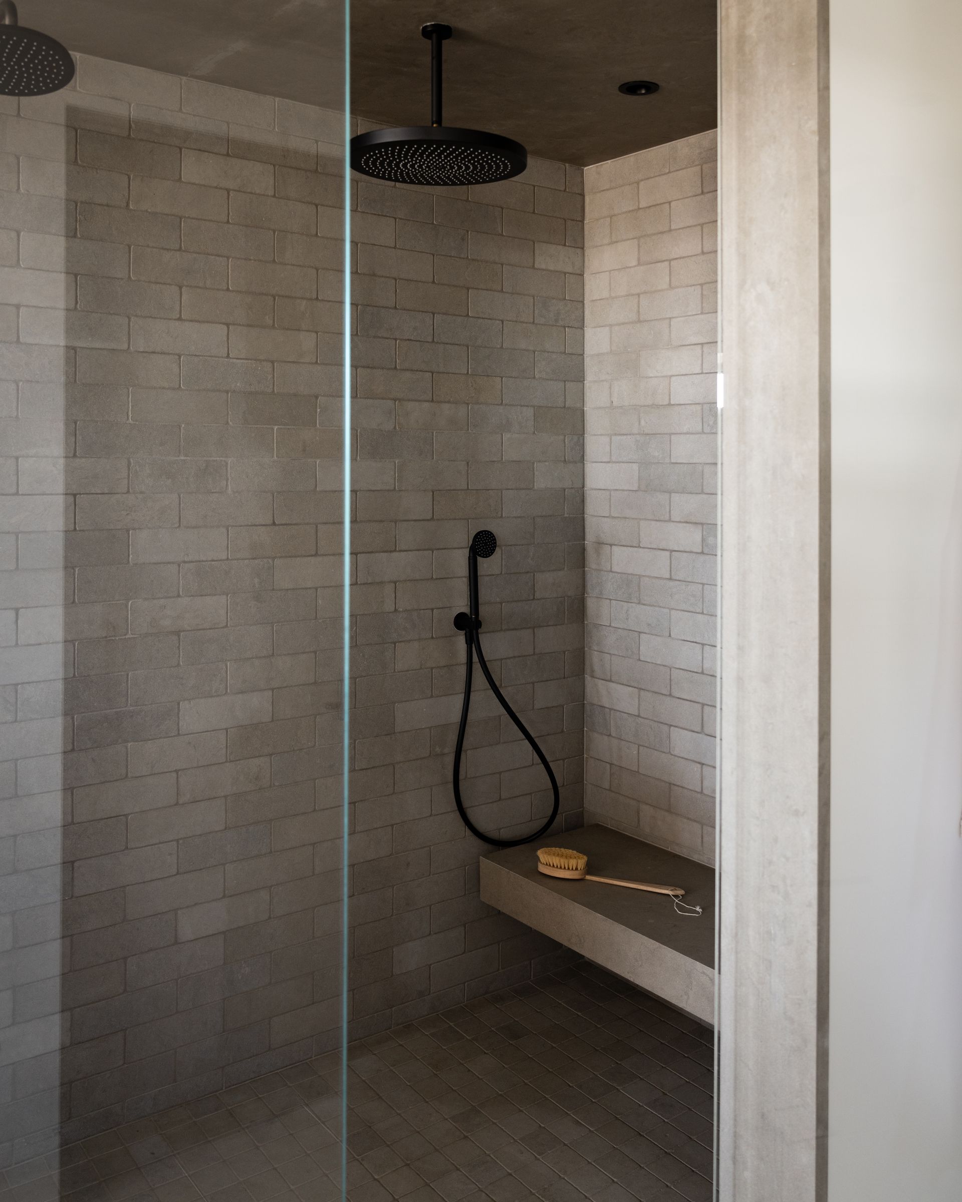 A modern walk-in shower with grey brick-style tiled walls, a black rainfall showerhead, handheld sprayer, and stone bench.