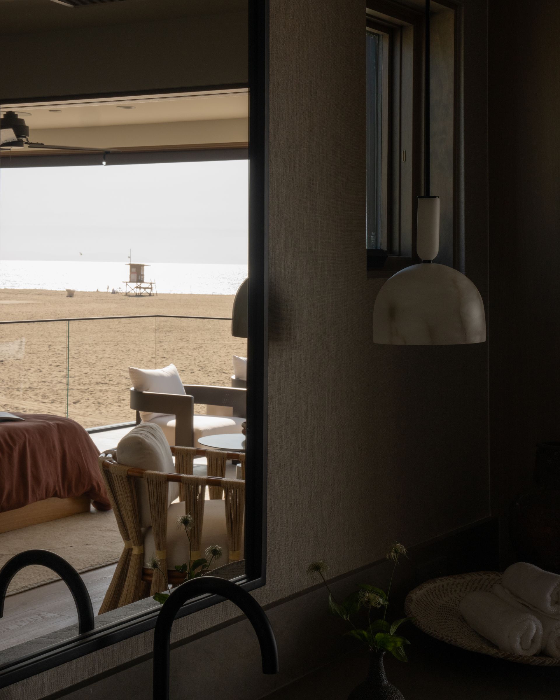 View from a bathroom through a large window overlooking a sandy beach with lounge chairs and a lifeguard tower.