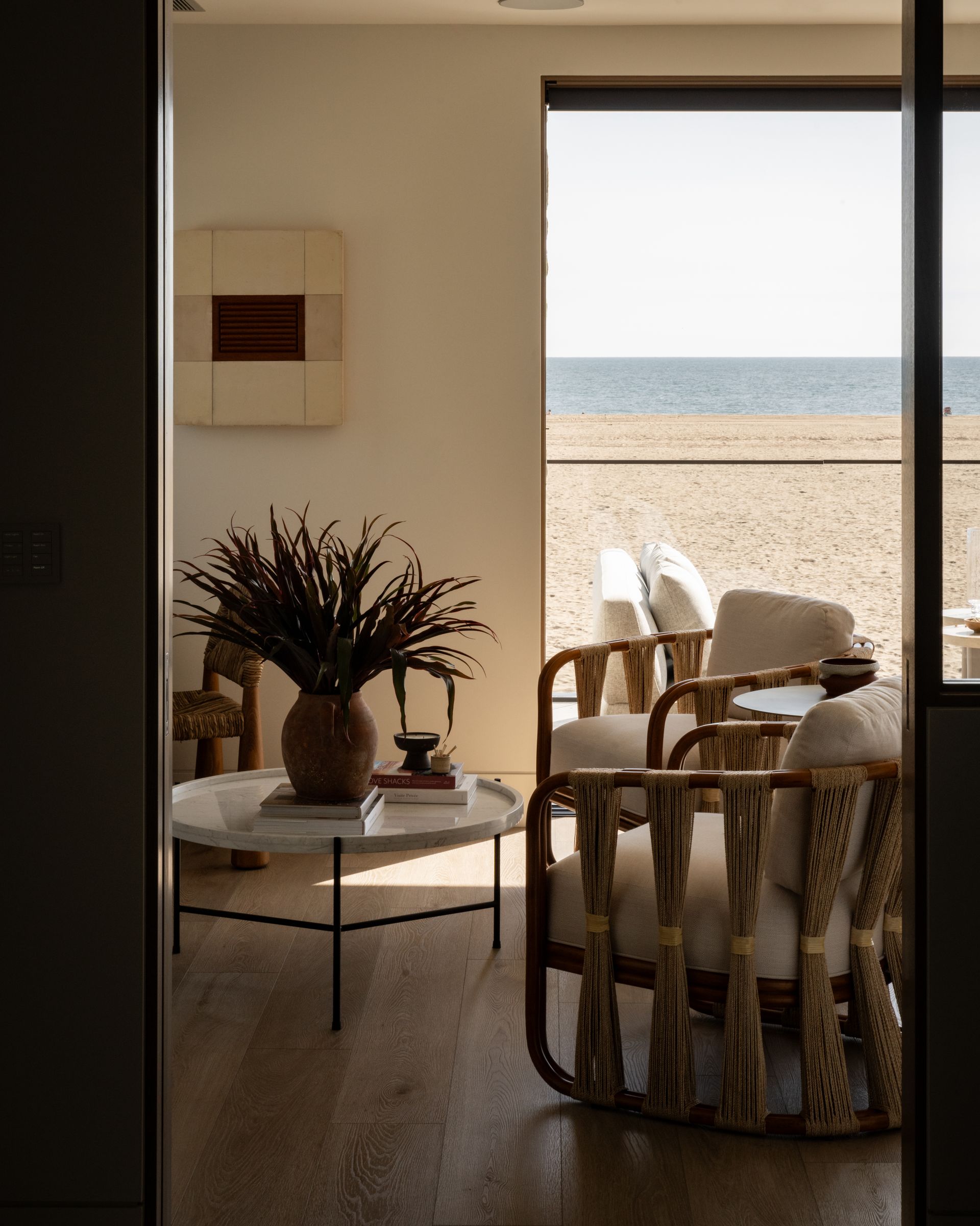 A sunlit room with woven chairs and a round coffee table, overlooking a sandy beach and the ocean.