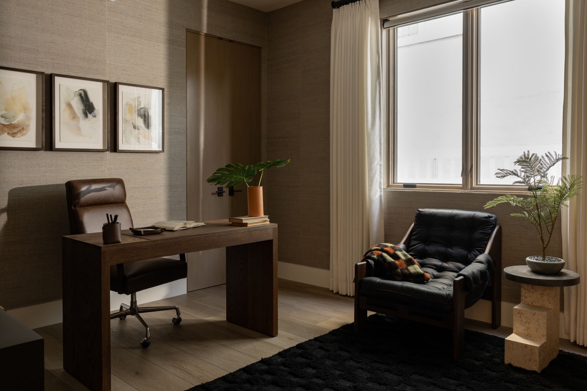 A modern office featuring a wooden desk, leather chair, and black armchair on a rug, with artwork and plants in the room.