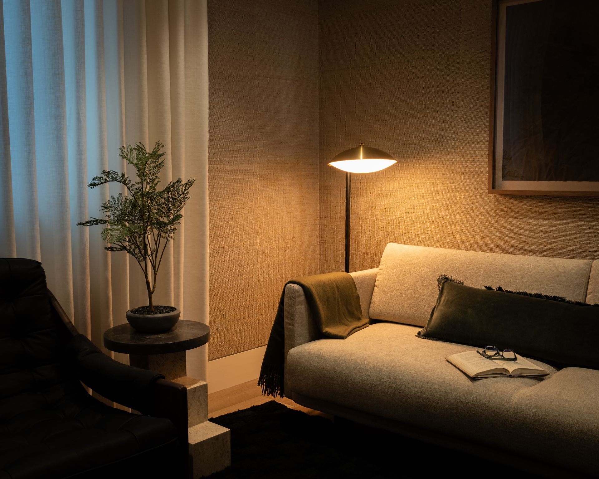 A cozy, dimly lit room with a beige sofa, a floor lamp, a small potted plant on a side table, and a black armchair.