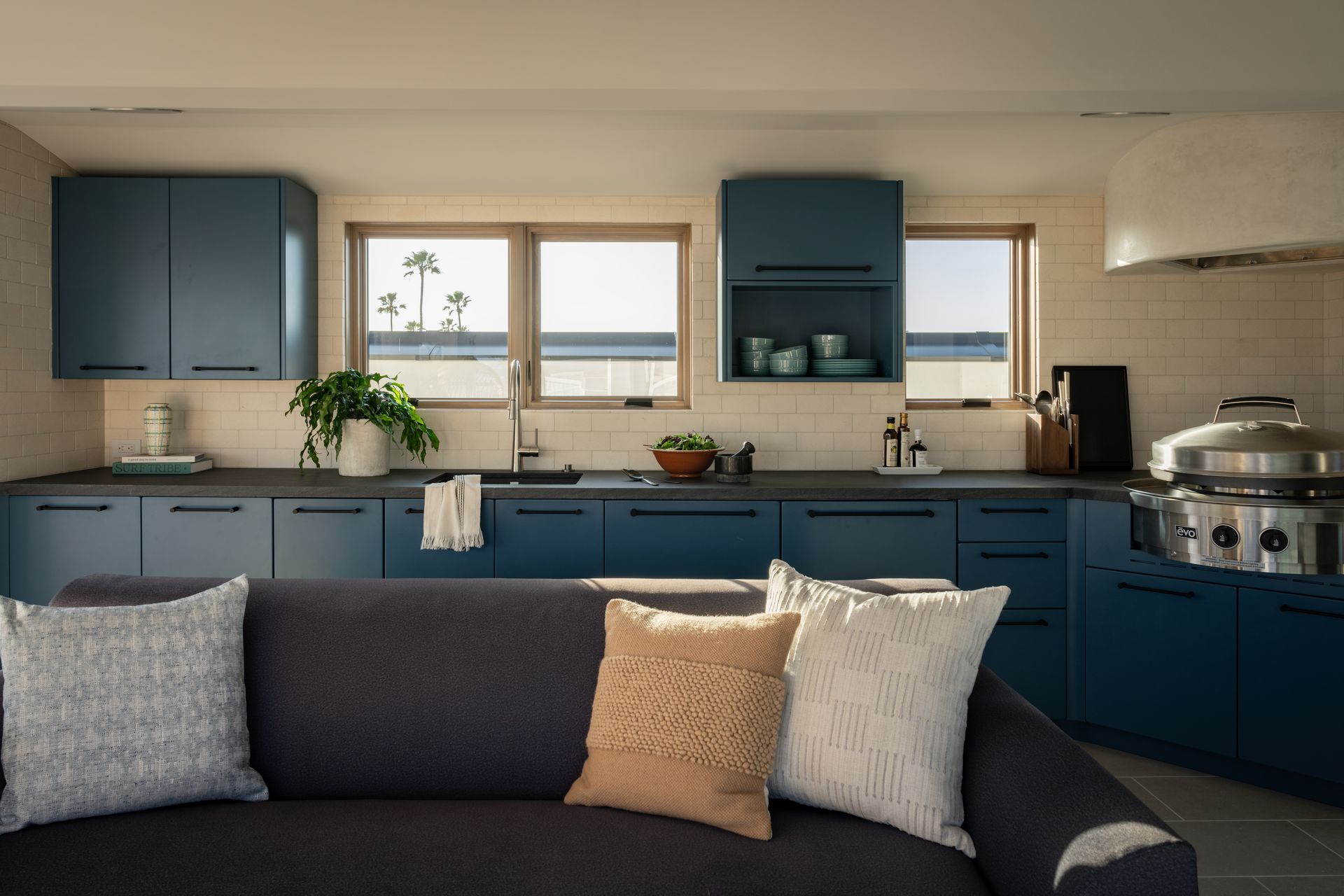 A dark blue kitchen with a couch in the foreground, featuring blue cabinets, light-colored countertops, and two windows.