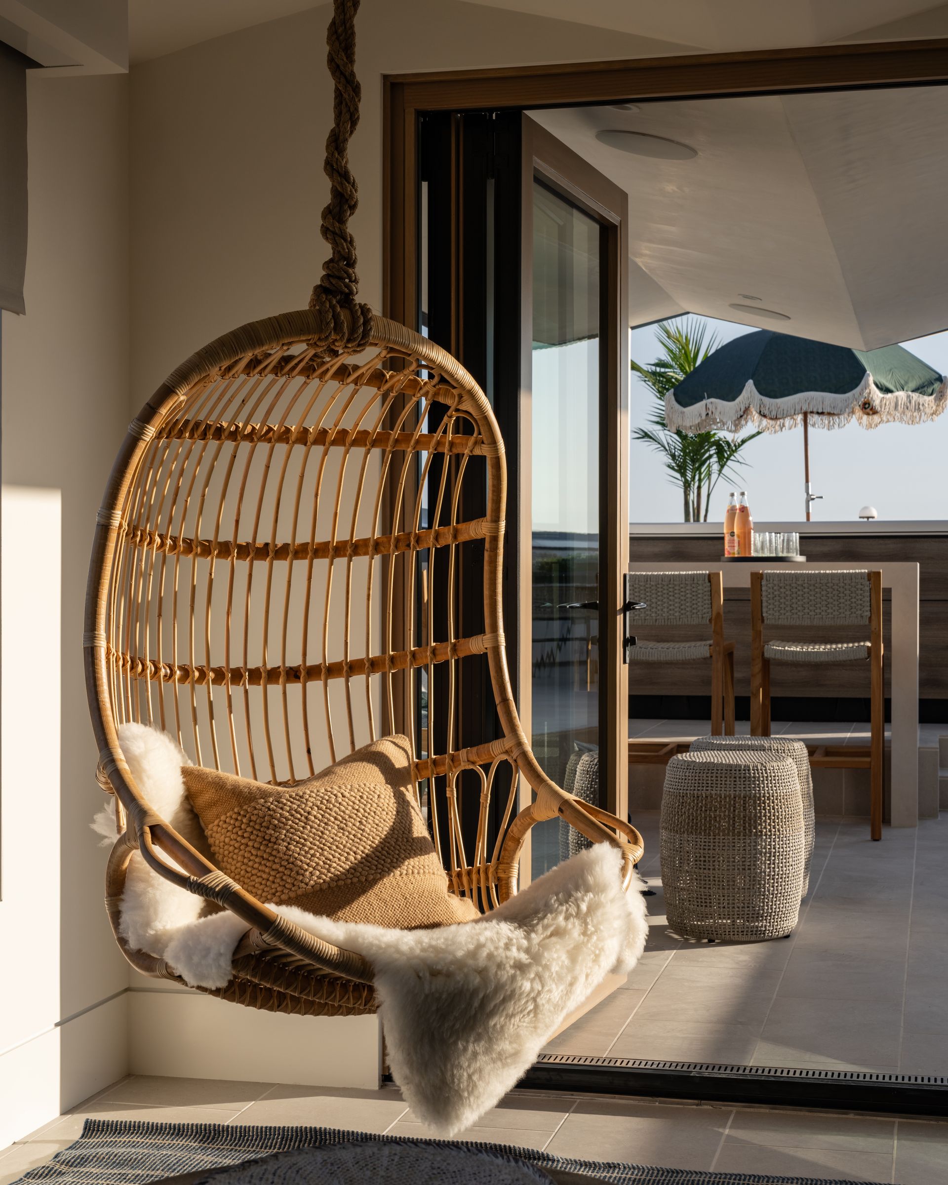 A hanging rattan egg chair with faux fur cushions sits indoors, overlooking a sunny patio with a green umbrella.